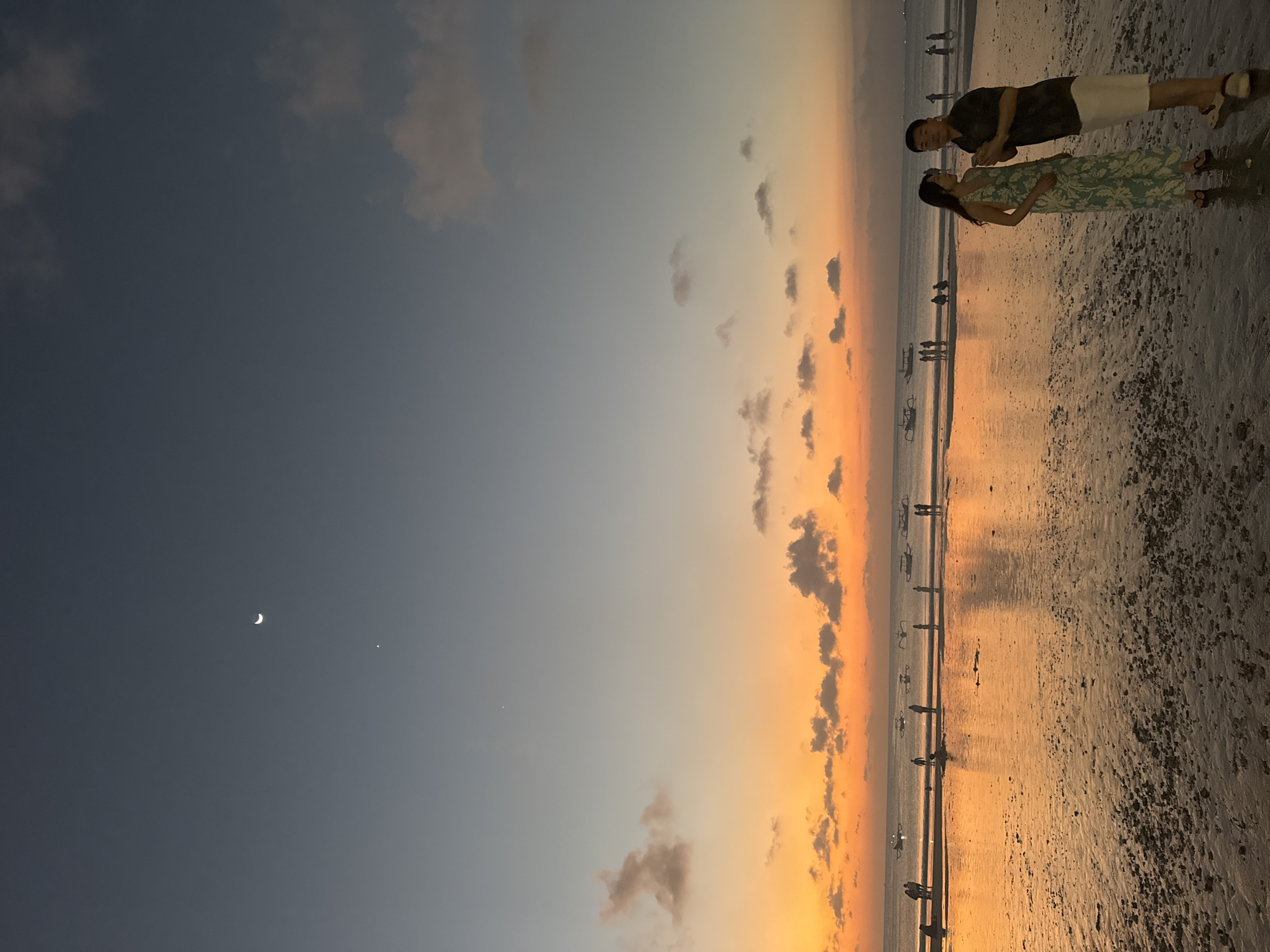 This is a full-frame, eye-level, vertical photograph of a beach scene in Kuta Selatan, Indonesia, during twilight. The sky transitions from a pale blue at the top to vibrant orange and yellow hues near the horizon, with scattered clouds. A crescent moon and a bright planet are visible in the upper portion of the sky. The sea is calm and reflects the colors of the sky. Several small boats are dotted across the water. On the wet sand, a group of people are standing and walking. In the foreground, a man and a woman are standing and facing each other, appearing to be in conversation. The man wears a dark short-sleeved shirt and white shorts, while the woman is in a long, patterned dress. The beach is composed of wet sand with some pebbles and debris.
