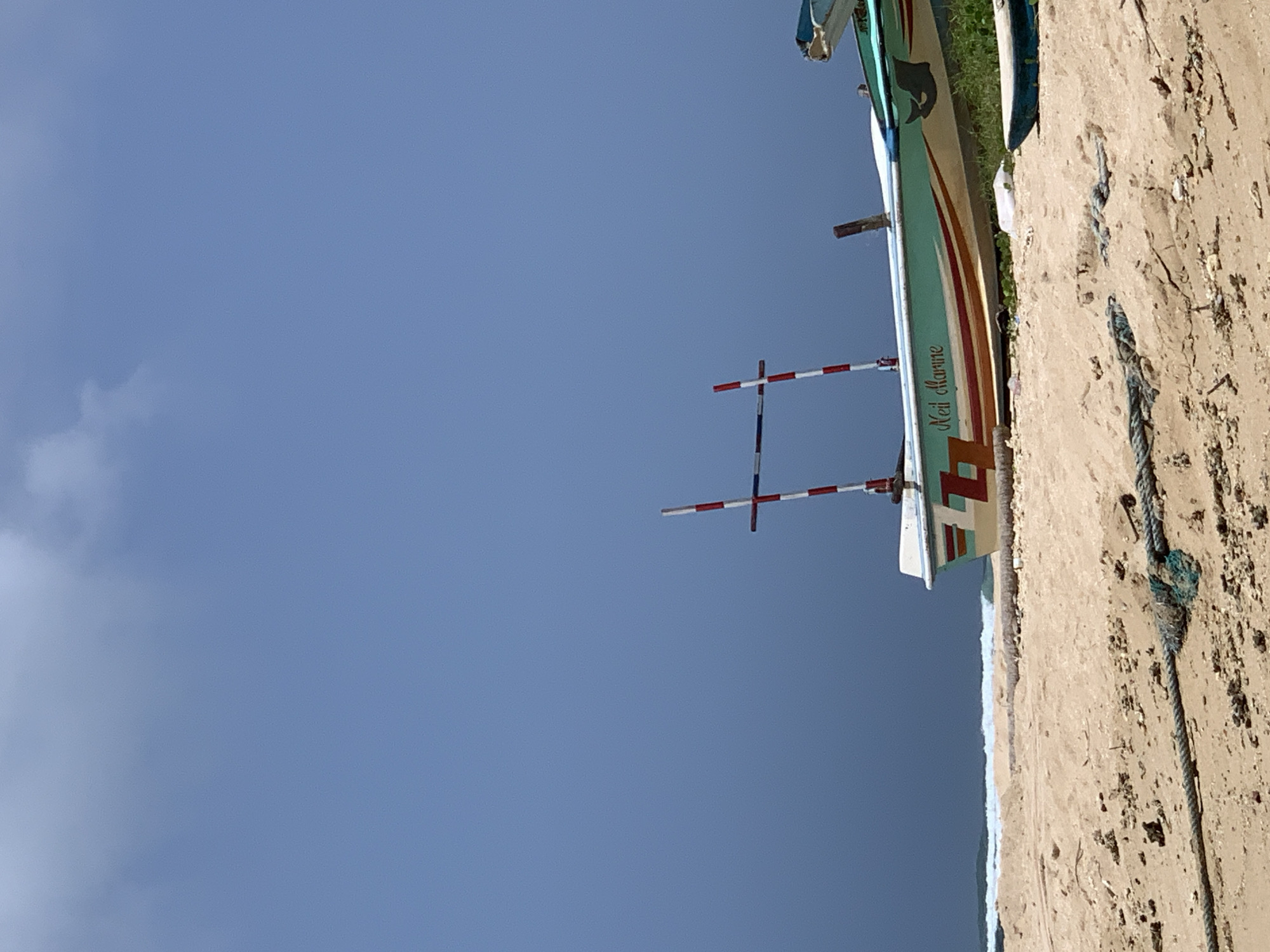 The image depicts a beach scene in Weligama, Sri Lanka, under a cloudy sky. In the foreground, a sandy beach is covered with debris and a thick rope. In the midground, a small fishing boat with the name "Neil Marine" painted on its side rests on the sand. The boat is decorated with red, white, and blue stripes. Another smaller boat is visible to the right of the main boat. In the background, the ocean meets the shore with gentle waves. The overall atmosphere is tranquil and serene, suggesting a quiet day on the coast.