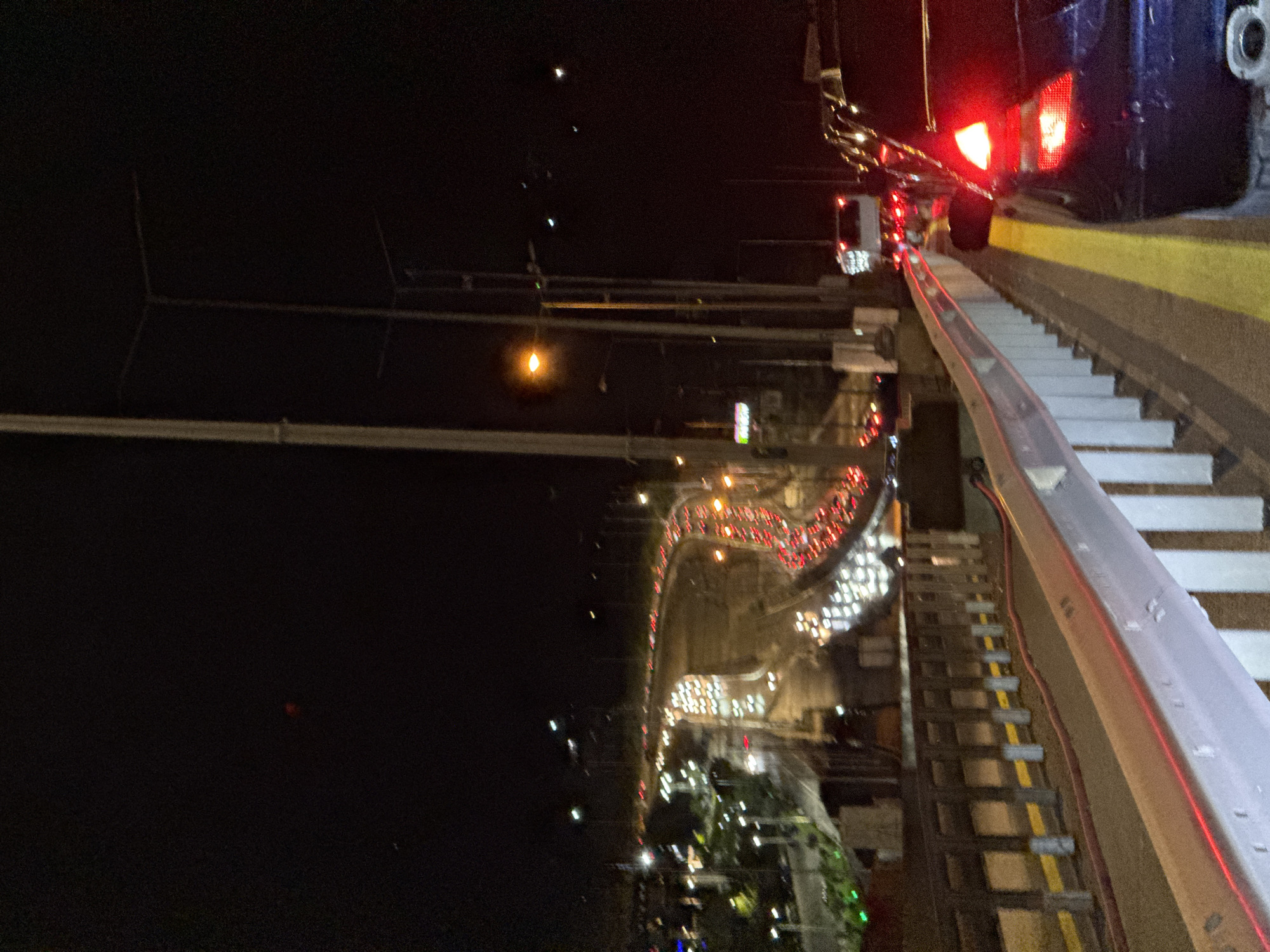 The image shows an elevated view at night, seemingly from a walkway or bridge, overlooking a road with heavy traffic in Pylaia Municipal Unit, Greece. The vehicle tail lights appear as red streaks on a curved road, indicating slow-moving traffic or congestion. A street lamp is visible. The presence of a walkway or stairs suggests an urban environment. A vehicle is partially captured on the right side of the frame, possibly parked nearby. The overall environment is dimly lit, with limited visibility beyond the immediate area of traffic and street lighting. There are no visible people in the image.