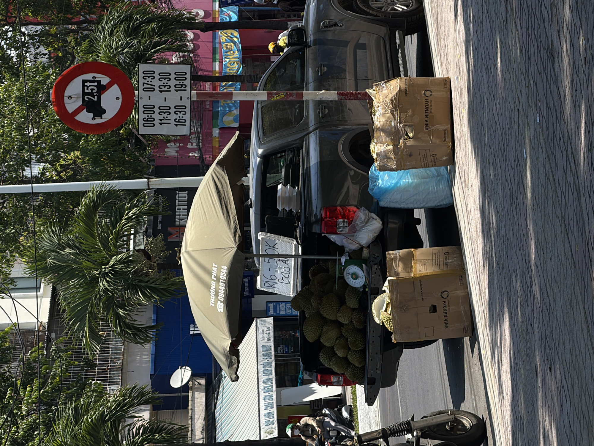 A grey pickup truck is parked on the side of a city street, likely in Nha Trang Ward, Vietnam, laden with durians. The truck bed is full of the spiky fruit, with some already cut open. A manual scale is placed on the truck bed, suggesting the fruit is for sale. A green umbrella is extended over the truck bed, providing shade for the durians and a sign with Vietnamese text and prices. There are also several cardboard boxes and blue plastic bags next to the truck. In the background, a traffic sign restricts vehicles over 2.5 tons during specific hours. Palm trees line the street, and buildings with shops and windows are visible behind them. A person on a motorbike is seen in the left foreground, and another person in a yellow vest is visible in the driver's seat of the truck. The street is paved with brick-like tiles, and sunlight casts shadows.