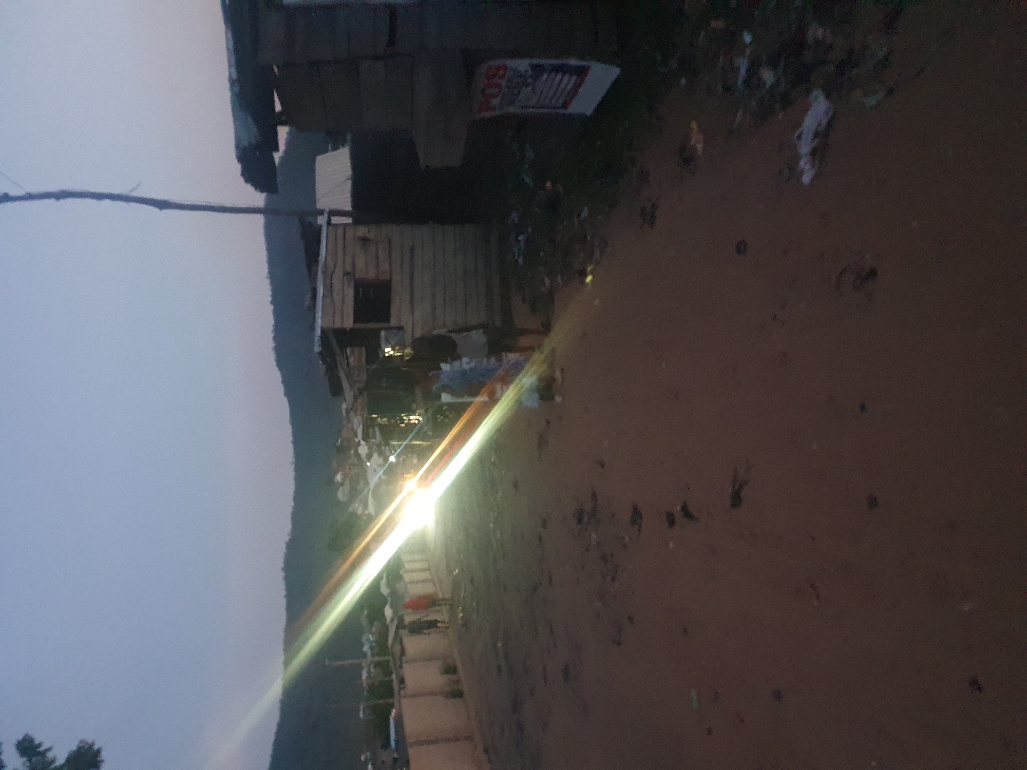 The image depicts an outdoor scene at dusk, likely in a low-income area of Lokoja, Nigeria. A dirt road runs along the right side of the image. Adjacent to the road is a small, dilapidated structure built from wood and possibly cinder blocks. Debris and trash are scattered around the structure. A hillside is visible in the background on the left. Power lines or a similar utility line runs across the top left. A bright light, possibly a streetlight or headlamps of a vehicle, creates lens flare across the center of the image. The overall impression is one of poverty and environmental degradation. No individuals are visible.