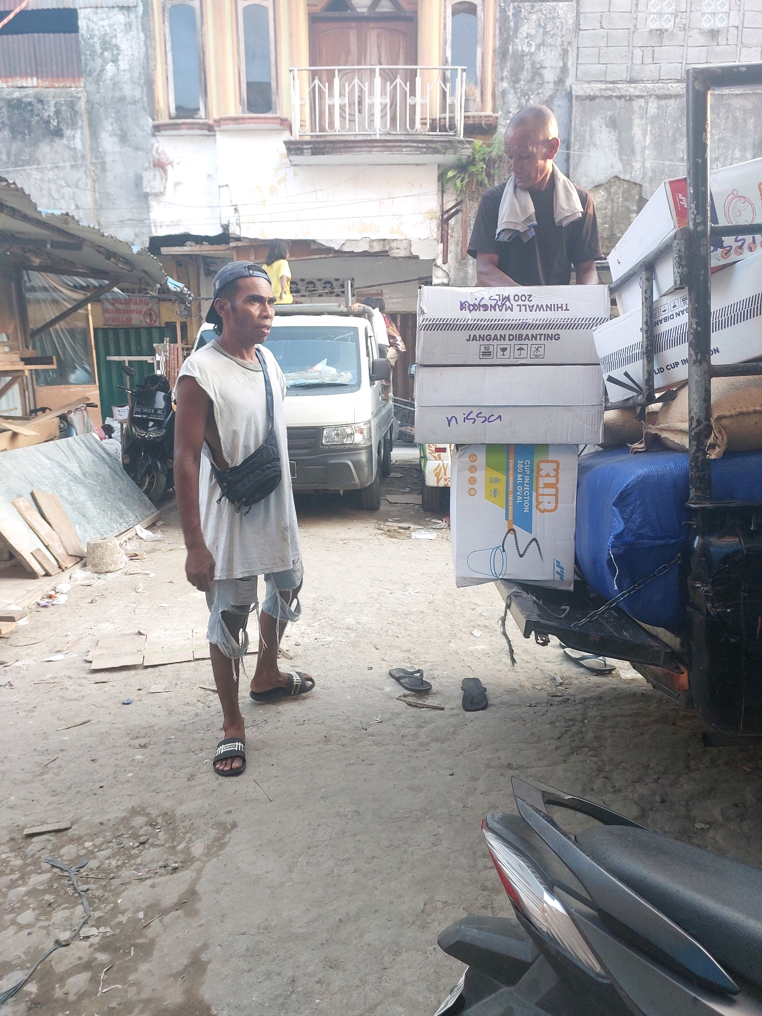 An eye-level, medium shot captures a man standing in a dusty urban alley in Ambon, Indonesia. The man, wearing a sleeveless white shirt, ripped denim shorts, and a blue baseball cap, faces right with his mouth slightly open as if speaking. A black crossbody bag is slung over his left shoulder. He stands near a white truck laden with cardboard boxes and some burlap sacks. To the right, another man, wearing a black t-shirt and a towel draped around his neck, is partially obscured by the boxes. In the background, a multi-story building with peeling paint and balconies stands prominently. The ground is a mixture of dirt, gravel, and scattered debris. A black motorcycle is visible to the left of the truck. The lighting suggests it is daytime, with a slightly hazy quality.