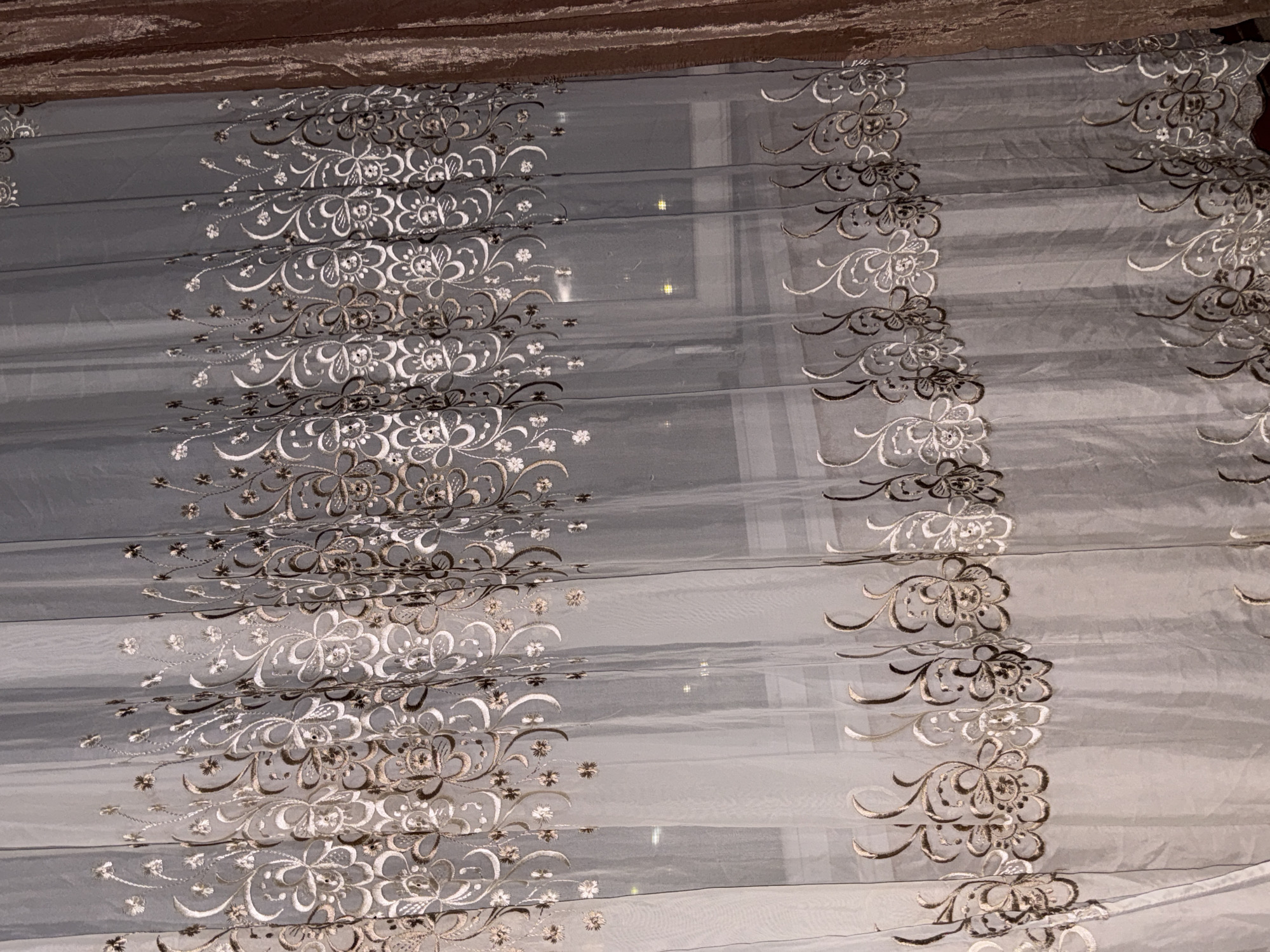 The image shows a close-up of a sheer curtain with an embroidered floral pattern. The curtain is a light, translucent fabric, and the embroidery features delicate swirls and flower motifs in shades of beige and white. The curtain is hanging vertically, and several vertical pleats are visible. In the background, a window is partially obscured by the curtain, and reflections of lights can be seen on the glass, suggesting it is nighttime or a dimly lit room. To the right of the sheer curtain, a darker, thicker curtain, possibly made of velvet, is visible. The image does not provide any information about the location in the city of Cahul, Moldova.