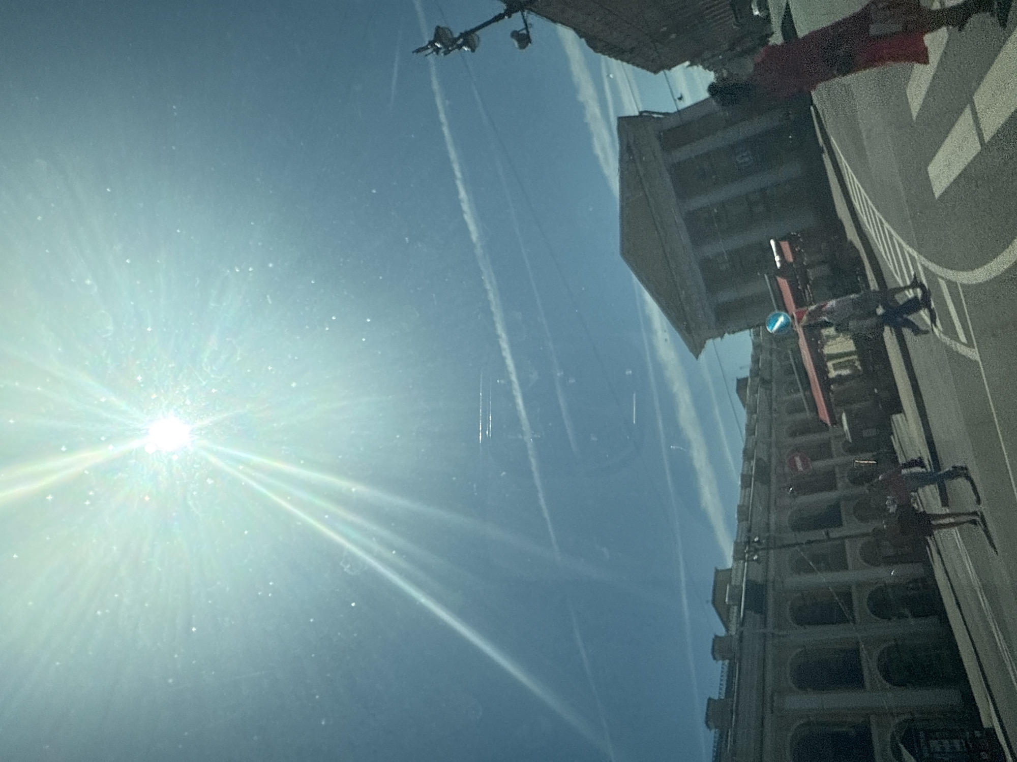 This is a sun-drenched, slightly overexposed exterior shot of a city street, likely Saint Petersburg, Russia, given the architectural style of the buildings. The sun, high in the bright blue sky, creates a strong glare and lens flare, with prominent light rays emanating from it. Several faint contrails streak across the sky.
In the foreground, a road with white pedestrian crossing lines curves towards the right. A person in a red dress is walking across the street from right to left. To the left of the crossing, two individuals, one in a light-colored top and dark pants, the other in a darker top and light pants, are walking across the street from left to right. Further to the left, two more individuals are walking away from the camera on the sidewalk.
Dominating the mid-ground is a large, imposing building with a classical facade, featuring columns and an arched entrance. To its left, a long, low building with numerous arched windows stretches across the scene, its exterior appearing weathered and possibly historic. A red awning extends from this building. On the far right, a tall, ornate lamppost with multiple lights is visible, partially silhouetted against the sky. The overall impression is a sunny day in a European city with notable historical architecture.