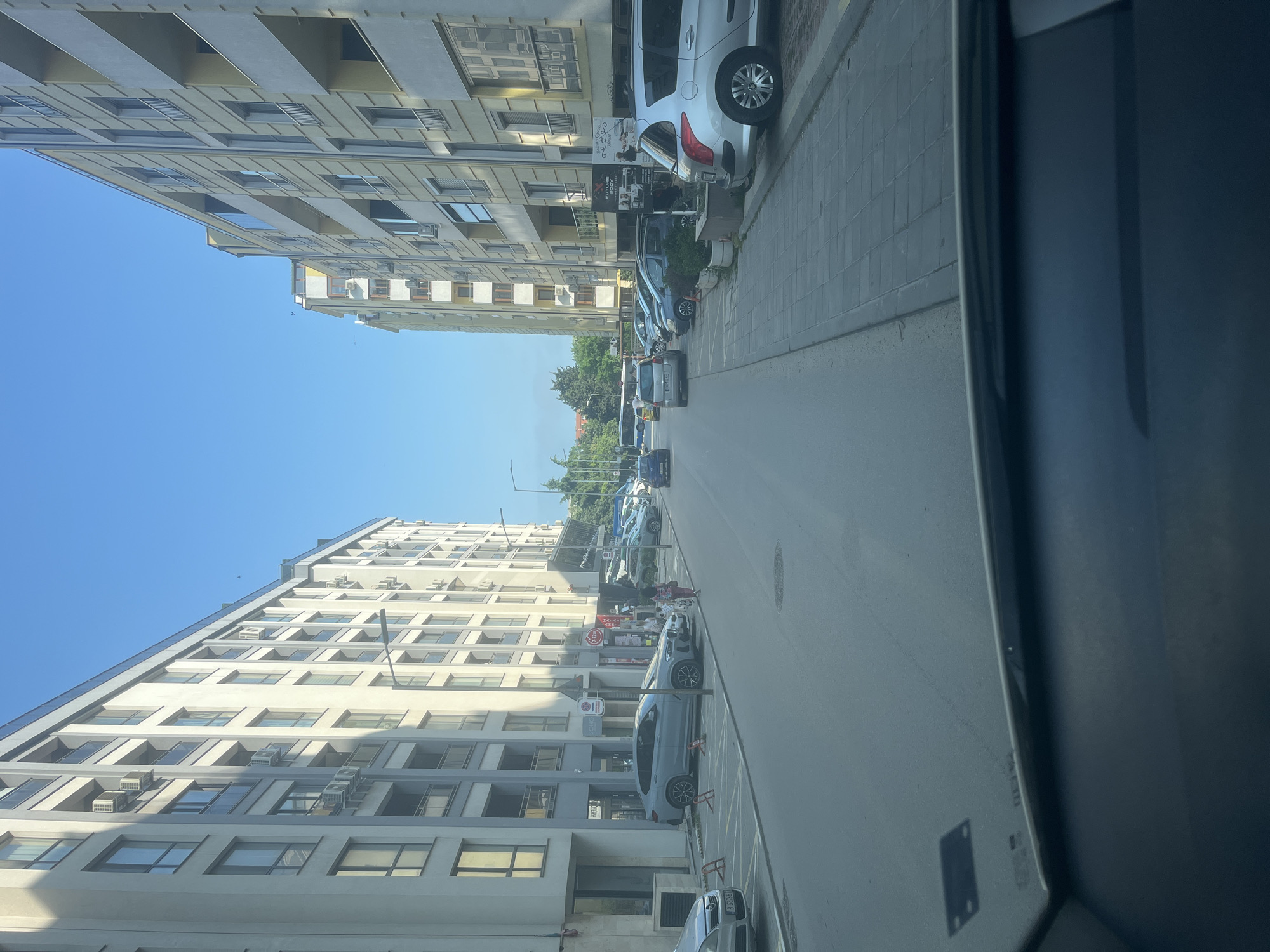 This image captures a bright, sunny daytime street scene in an urban environment, likely in Varna, Bulgaria, as indicated by location cues. The photo appears to be taken from inside a vehicle, with a dark frame or dashboard visible in the foreground.
The scene features a wide street lined on both sides by multi-story buildings. On the left, a tall, modern building with many windows and balconies dominates the view, its light facade reflecting the clear blue sky. On the right, older-style buildings with varied architectural details and ground-floor businesses line the sidewalk.
Numerous cars are parked along both sides of the street, some partially on the paved sidewalk. Vehicles visible include a silver hatchback (possibly a Peugeot 308), a light blue SUV, and other sedans. The road in the center is clear, suggesting active traffic or recent movement. In the distance, trees and green foliage indicate a park or landscaped area.
Notable details include the clear blue sky, suggesting good weather, and strong sunlight casting shadows. A red circular stop sign with the Cyrillic text "СТОП" (STOP) is visible on the right. Storefront signs include "FLOWERS" and "BEAUTY SALON" on a vertical sign, and "SOLAR" on another. A Bulgarian license plate starting with "B 5..." is faintly visible on one of the parked cars, with "B" being the district code for Varna. The overall atmosphere is that of a typical, calm urban street. No specific event or strong emotions are evident.