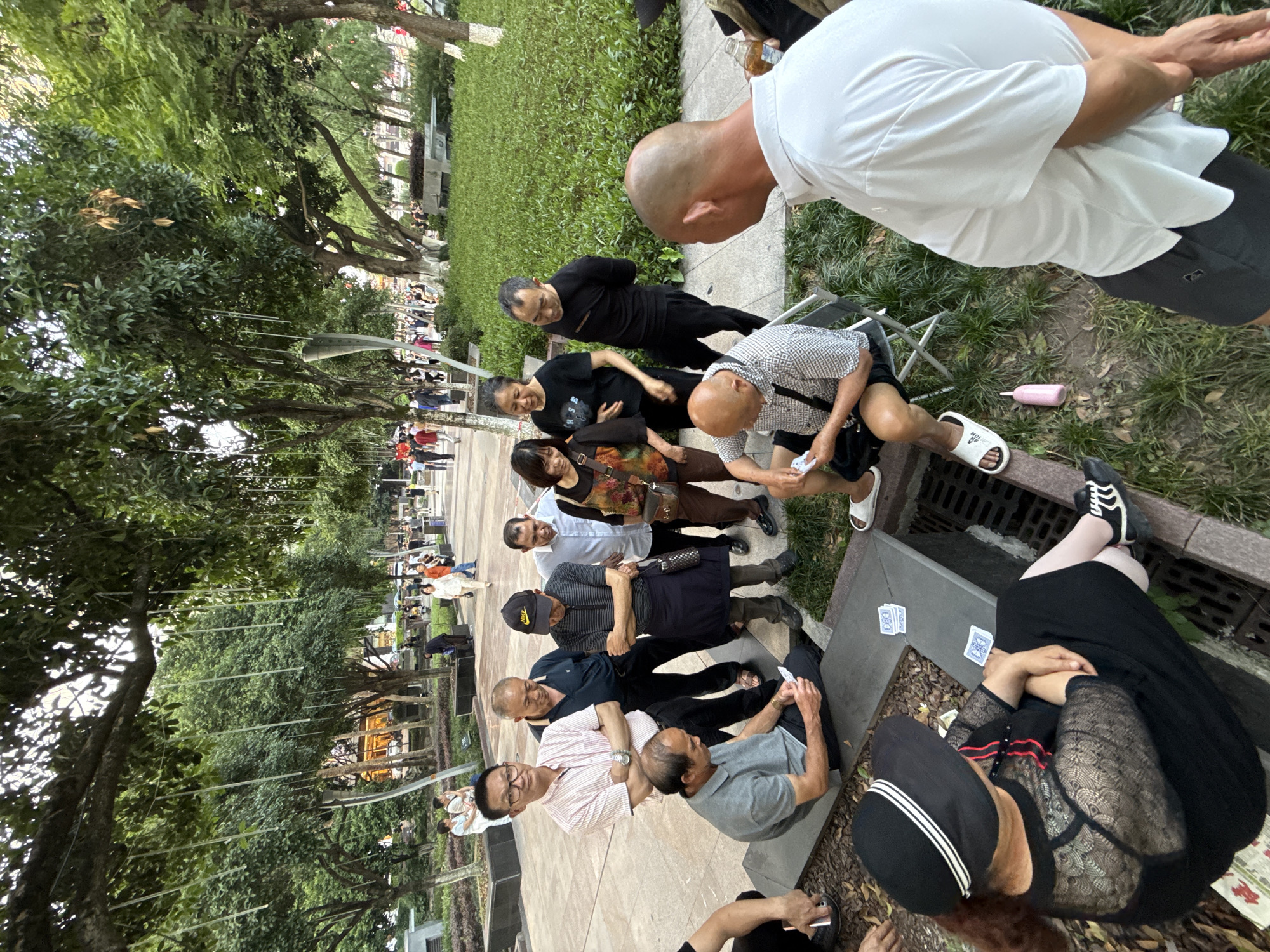A group of approximately ten people are gathered in a public outdoor space, likely a park, in what is reportedly the city of Liangjiang, China. The group appears to be middle-aged to elderly adults. Several are seated on a low stone barrier and along the edge of a concrete walkway, while others are standing. Some members of the group are interacting with playing cards, which are visible both in their hands and scattered on the ground nearby. The environment consists of trees, grass, and a paved area. People in the distance are partially visible along the periphery of the park.