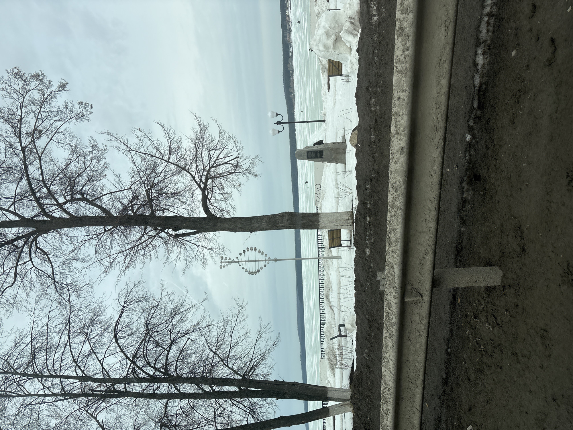 The image depicts a waterfront scene in Votkinsk, Russia. Bare trees frame a partially frozen body of water, possibly the Votkinsk Reservoir. Along the shore, snow covers the ground, punctuated by a few benches and a monument. A decorative metal sculpture stands near the waterfront. A guardrail runs along the foreground, suggesting the scene is viewed from a road or elevated pathway. The sky is overcast. There are no people visible.