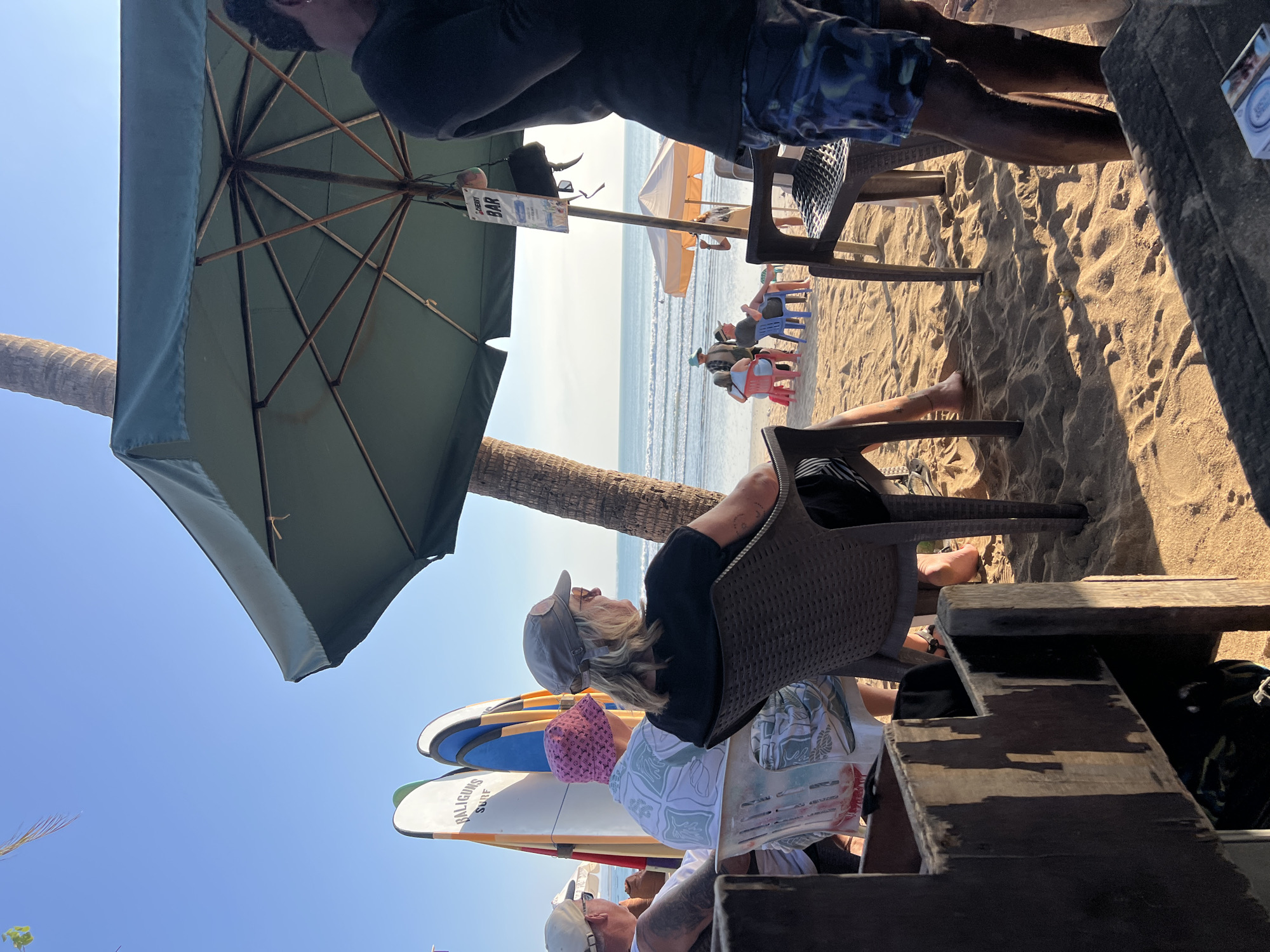 This is a photograph taken at a beachfront establishment in Kuta, Indonesia. The image captures a sunny day with a clear blue sky. In the foreground, to the left, there is a rustic wooden structure with two levels, appearing to be part of a seating area or bar. In the center, several people are seated on plastic chairs on the sand, facing the ocean. One person in the foreground on the right is standing and wearing patterned swim shorts.
Behind the seated individuals, a line of surfboards stands upright against a palm tree. The ocean is visible in the background with gentle waves and a few people in the water. A large green umbrella provides shade over some of the seating area. A sign for "CHERRY BAR" is hanging from a pole under the umbrella. The sand is a warm golden color, and the lighting suggests it is late afternoon or early morning, casting distinct shadows. The overall scene depicts a relaxed atmosphere at a beach bar or restaurant.