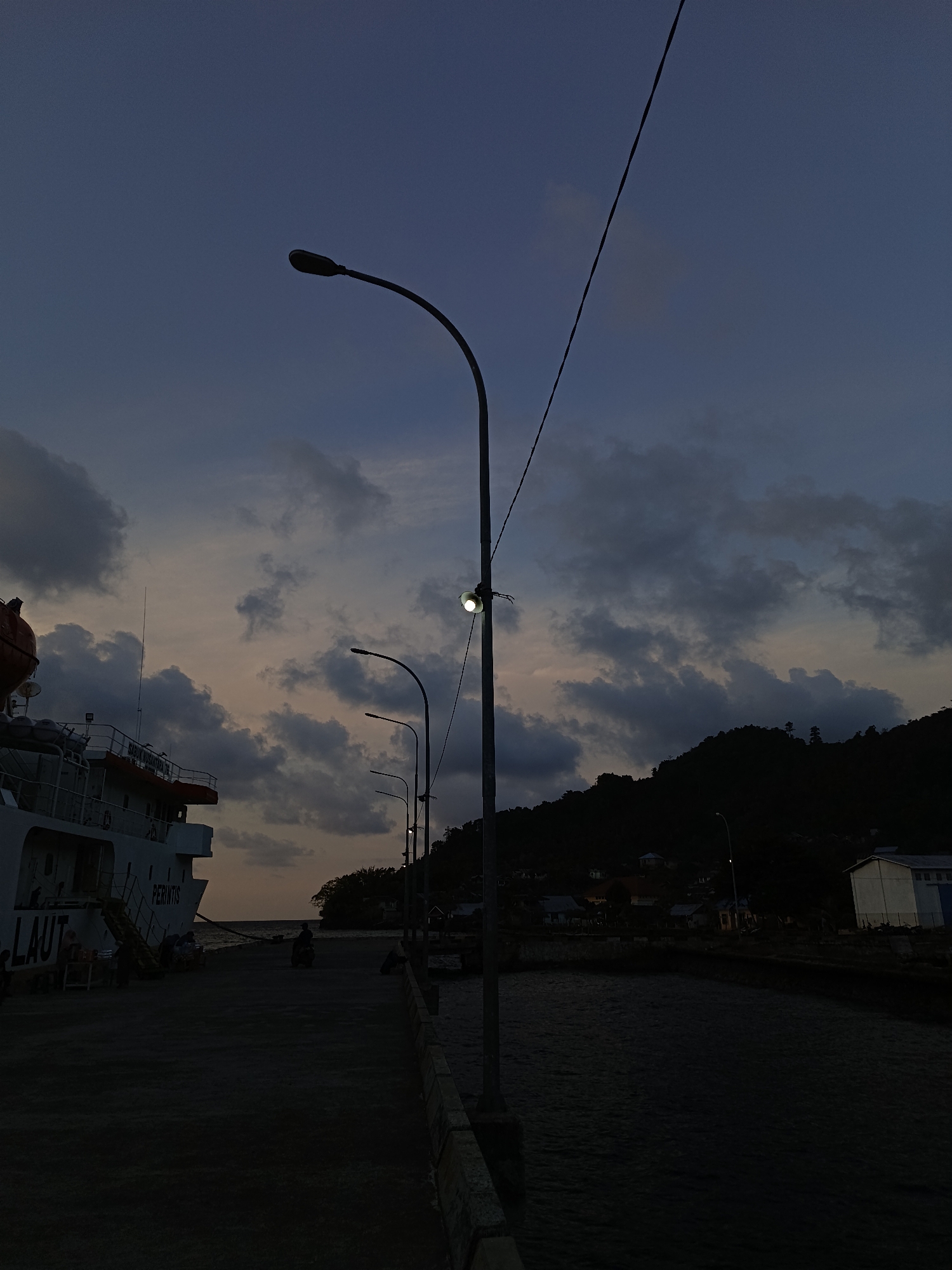 This image depicts a twilight scene at a harbor in Mansalean, Indonesia. A large, white cargo ship, identified with the partial text "LAUT PERINTIS", is docked on the left. The ship has an orange life raft visible at its stern. A concrete pier extends into the dark water. Several streetlights line the pier, with one prominently illuminated. In the background, a densely vegetated hillside with scattered houses rises, silhouetted against the dusky sky. Dark, heavy clouds dominate the upper portion of the frame, with a hint of soft light breaking through near the horizon. A person on a motorcycle is visible on the pier in the middle ground, moving away from the camera. Other indistinct figures are present near the ship. The overall atmosphere is one of a quiet, early evening at a working harbor.