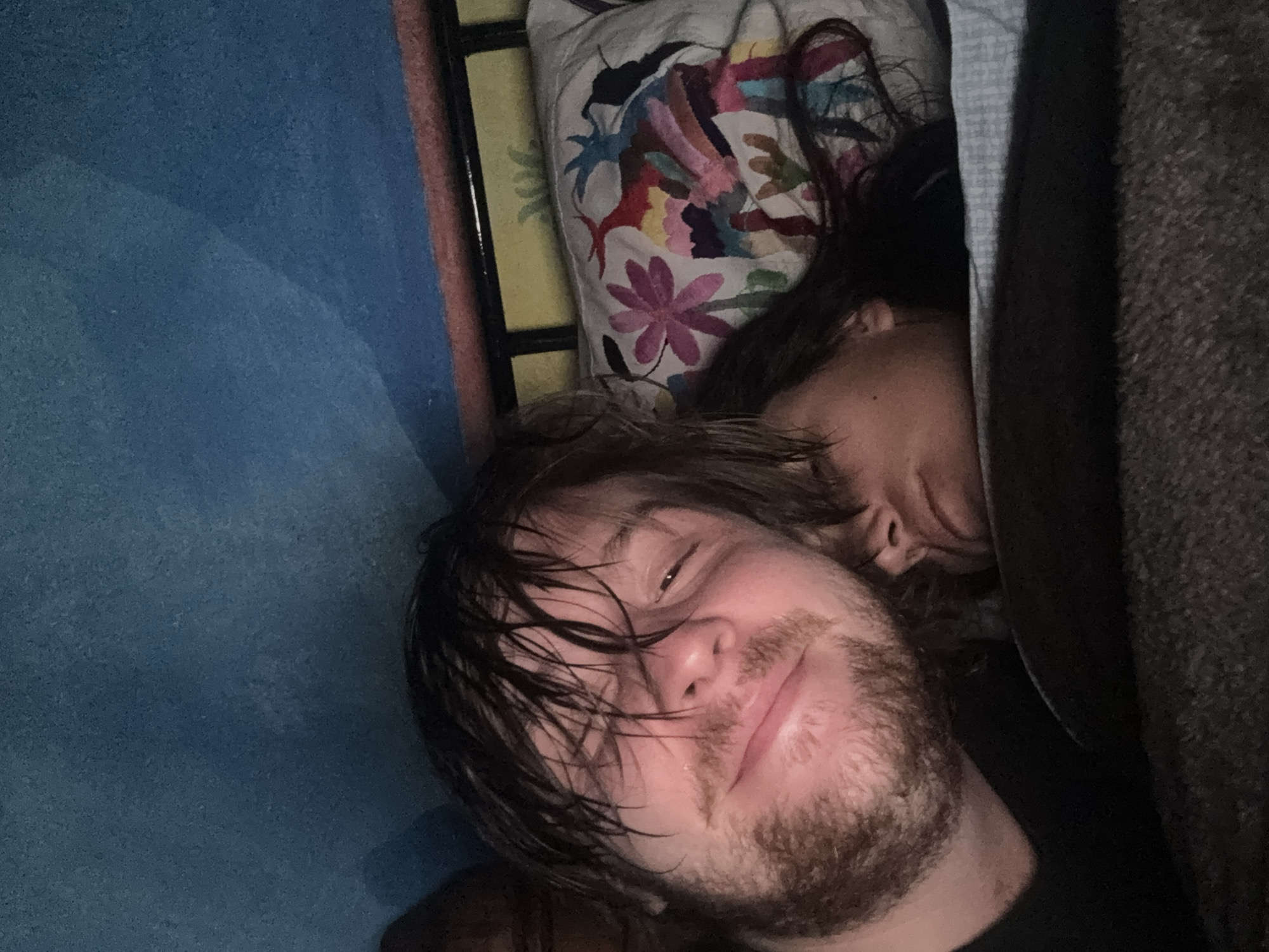 A close-up, dimly lit selfie captures a man with wet, dark blond hair and a beard, resting alongside a woman. The woman, positioned behind the man, has dark hair and appears to be asleep. They are lying on a bed with a colorful pillow featuring floral designs. A gray blanket covers part of the scene on the right side. A blue wall and a bed frame are visible on the left. The location, Cañada de Madero, Mexico, is not directly discernable from the visual information provided.