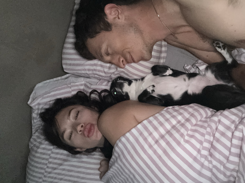 This image depicts an indoor bedroom setting in Tel-Aviv, Israel. Two adult individuals and a dog are positioned on a bed. On the left, a female individual with dark, wavy hair lies on her back, partially covered by a light-colored duvet with pink and white vertical stripes. Her left shoulder and upper arm are visible. Her face is directed towards the camera, with eyes partially closed and lips pursed in a 'kissing' or 'pout' expression. On the right, a male individual with short, dark hair and stubble lies on his left side, facing towards the female and the dog. He is bare-chested, wearing a thin necklace, and has his eyes closed or partially closed, exhibiting a subtle smile. Between the two individuals, a small to medium-sized black and white dog lies on its back, exposing its belly and paws. The dog's eyes are partially open and show a green reflection. The bedding, including pillows and duvet, features a consistent pattern of pink and white vertical stripes. The background beyond the bed is dark and indistinct, suggesting low-light conditions.