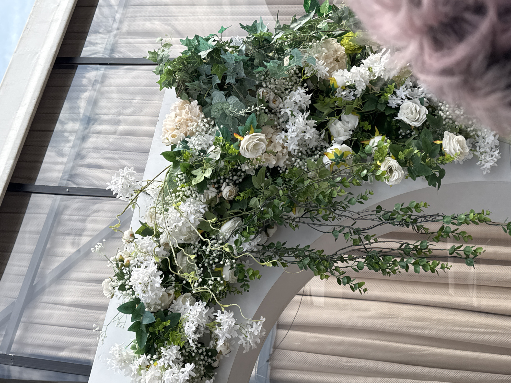 The image shows a close-up of a white floral archway decorated with white roses, hydrangeas, baby's breath, and lush green ivy. The archway is part of an outdoor event setup, with beige curtains visible in the background through a glass window. The sky and clouds are reflected in the glass. In the foreground, the blurry pink top of a person's head is visible, suggesting a celebration or event is taking place. This scene is located in Koy Tash, Kyrgyzstan.