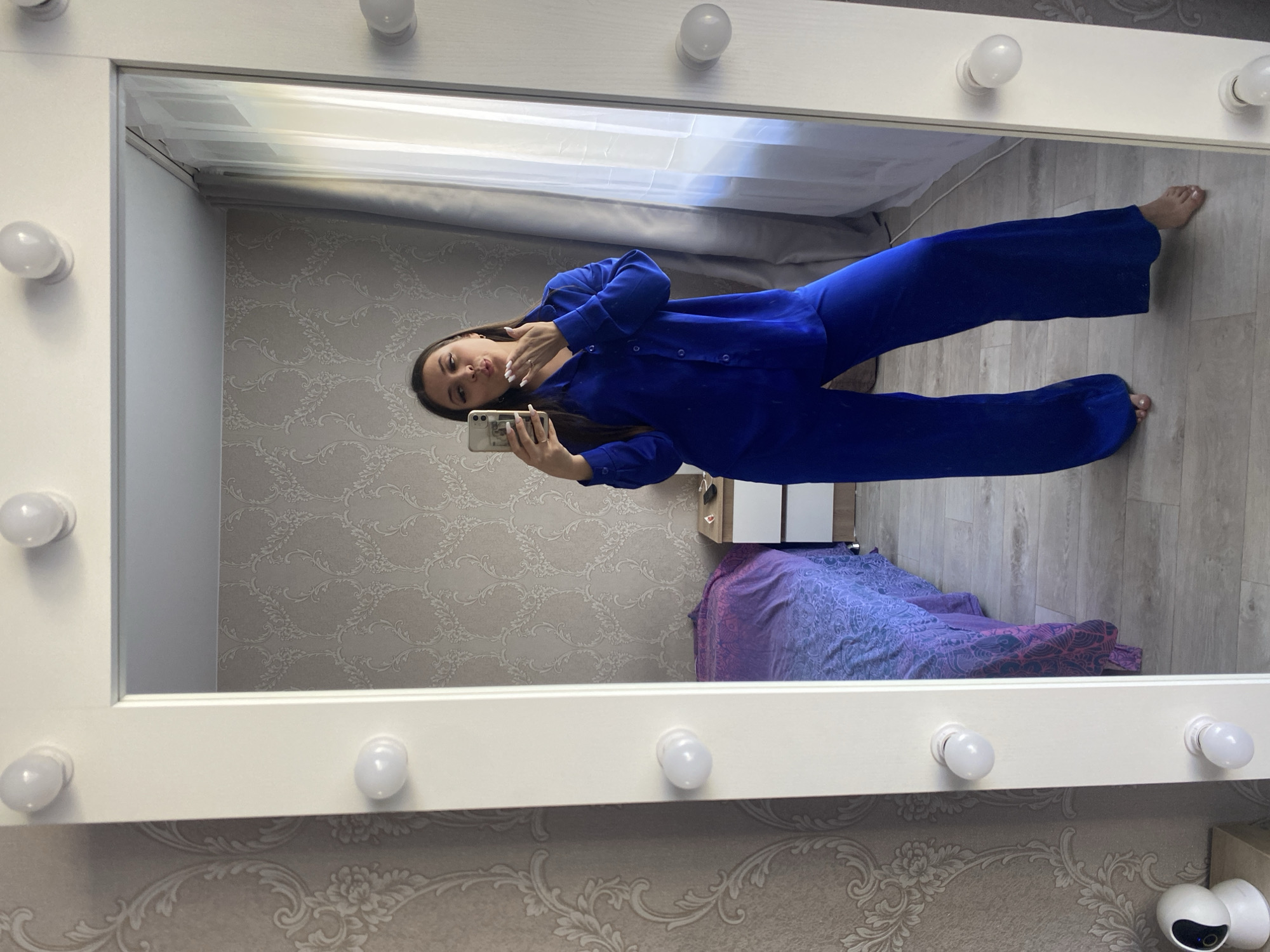 Here is a description of the image:
A medium shot depicts a mirror with light bulbs around the frame, reflecting a woman in a blue pajama set taking a selfie with her phone. The woman has long, dark hair and is posing with her hand near her face. The background shows a room with patterned wallpaper, a window with curtains, and a bed with a purple and blue patterned bedspread. A white cabinet is also visible in the reflection. There is no specific information in the image to confirm the location as Chaikovsky, Russia.