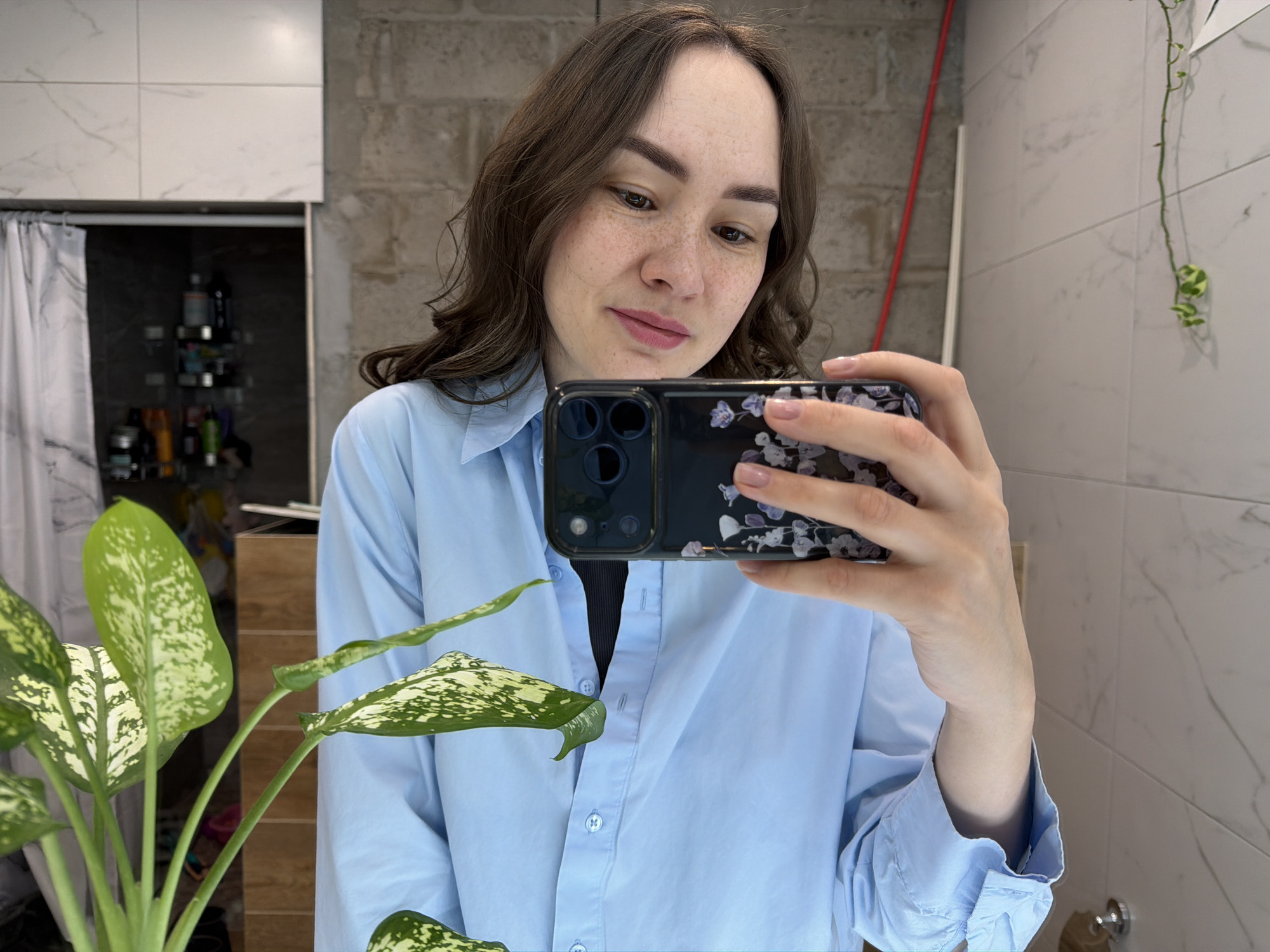 A woman is taking a selfie in a bathroom. She is wearing a light blue button-down shirt and holding a smartphone with a floral-patterned case. Her hair is brown and wavy. There is a plant with green and white speckled leaves on the left side of the frame. The bathroom has white marble-patterned tiles and gray brick accents. A shower with a curtain is visible in the background. A red pole is leaning against the wall. There are toiletries on a shelf inside the shower. A hanging plant is also visible on the right side of the frame. The location is Chaikovsky, Russia.
