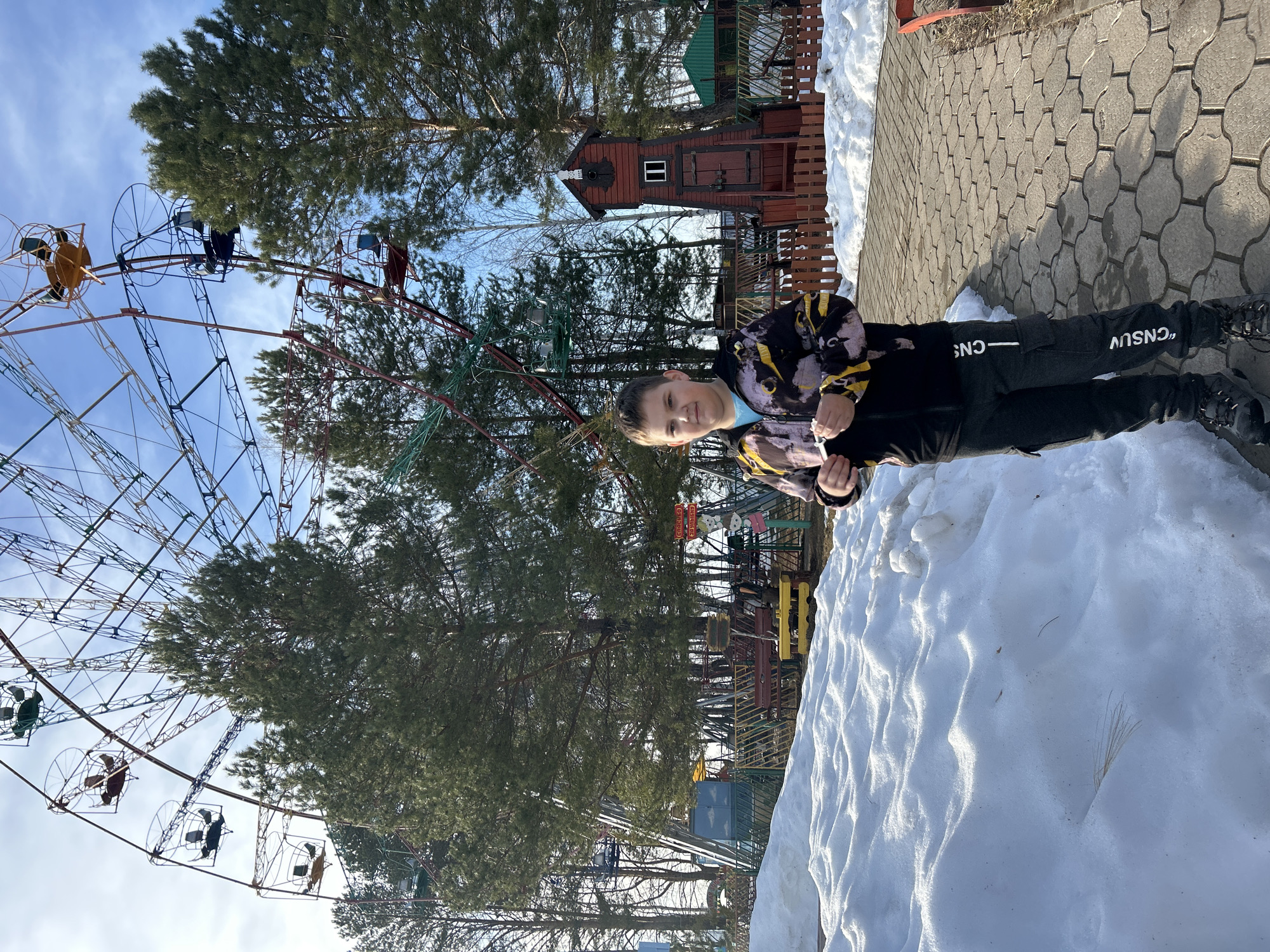 The image shows a child standing near a snowdrift, with a Ferris wheel and other amusement park rides visible in the background. The location is Chaikovsky, Russia. The child is wearing a black and yellow jacket and black pants with white writing. The snowdrift is large and covers part of the paved walkway. The Ferris wheel is multi-colored and appears to be an older model. Trees are interspersed throughout the amusement park area.