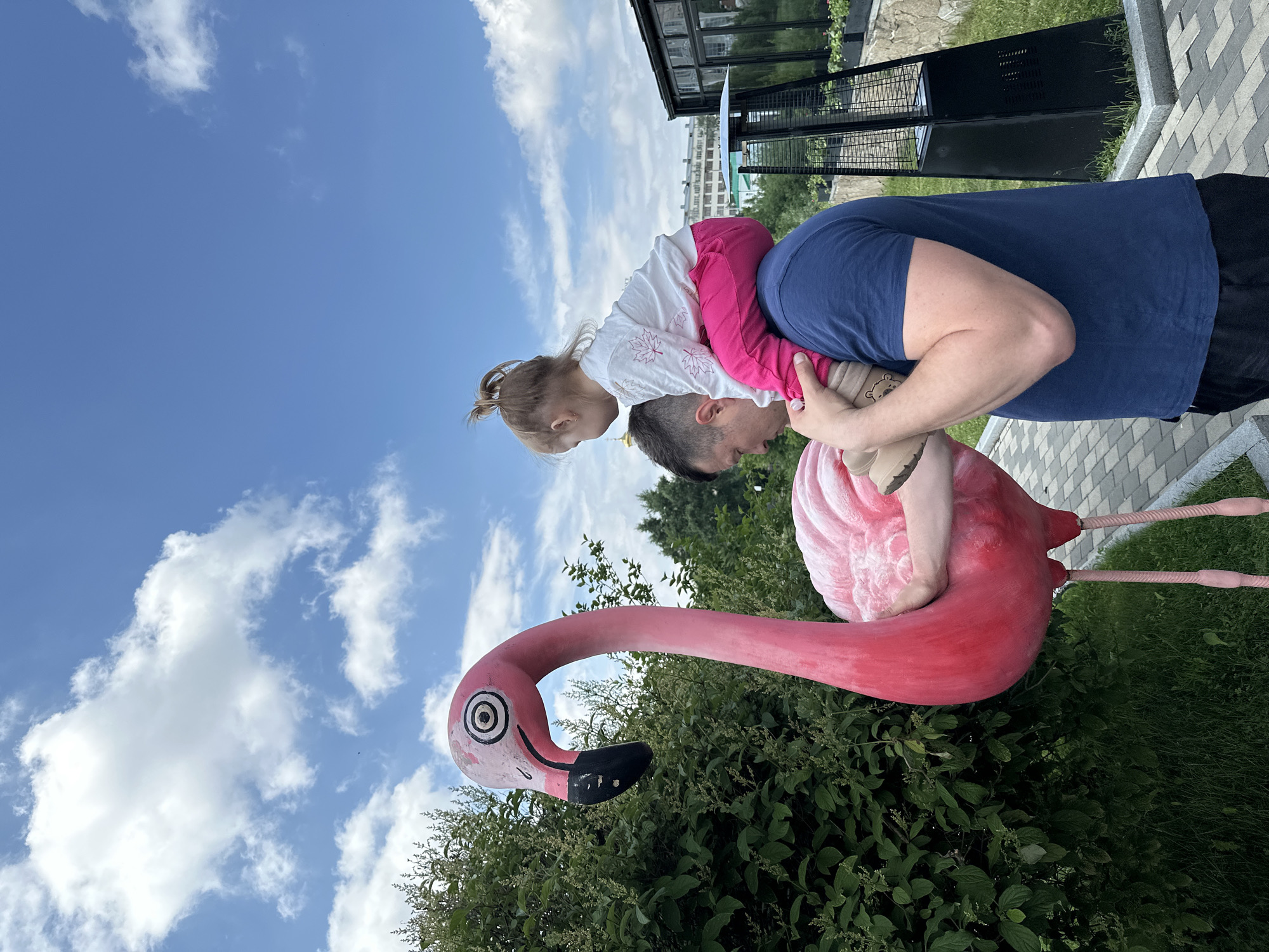 An outdoor, medium shot captures a man carrying a young child on his shoulders. The man, wearing a dark blue t-shirt, is facing to the left, his head tilted downwards. The child, dressed in a white long-sleeved shirt with pink leaf imprints and bright pink pants, faces away from the viewer, looking towards the left. They are positioned in front of a large, stylized pink flamingo statue. The flamingo is depicted with a black beak, large circular eyes with black pupils, and a textured body. Behind the flamingo and the pair, a dense green hedge forms the backdrop. In the background, a modern building with large glass windows and a tall, black outdoor heater are visible. The sky above is a bright blue, dotted with scattered white clouds. The ground is paved with gray and beige interlocking bricks. The image appears to be taken on a sunny day. The location is identified as the "flamingo park" in Yekaterinburg, Russia.
