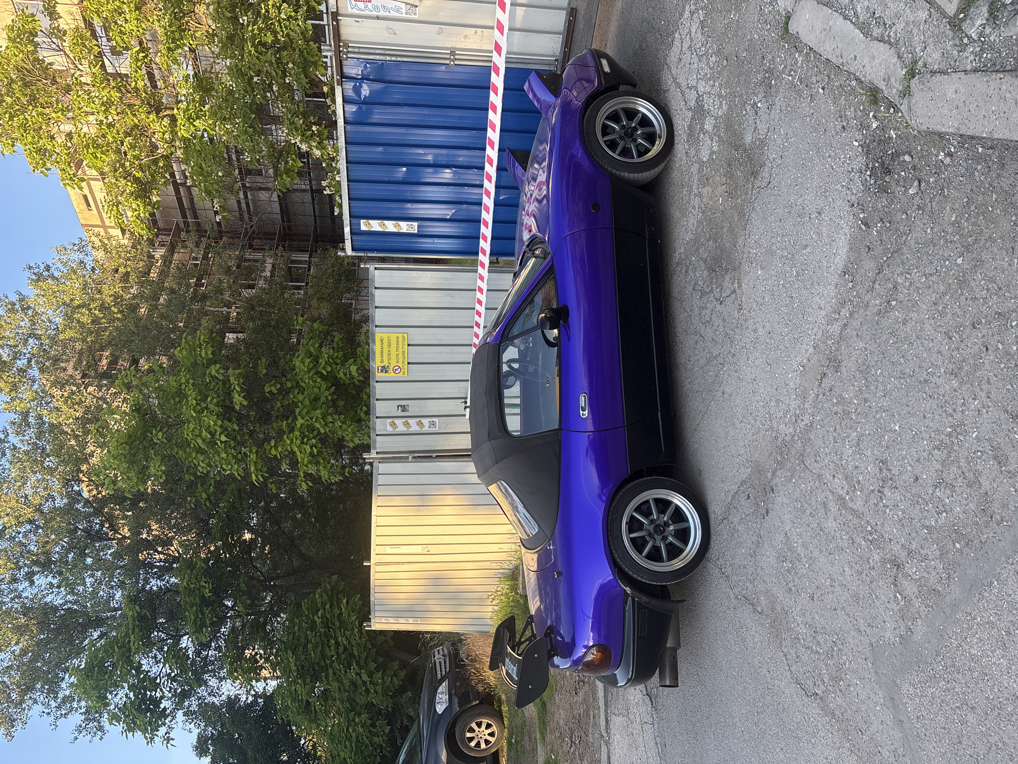 A purple and black two-door convertible sports car with a large rear spoiler is parked on a cracked asphalt surface. The car has a black soft top. To the left of the sports car, a black SUV is partially visible. Behind the vehicles, a white corrugated metal fence runs horizontally, with a blue corrugated metal fence segment to its right. A construction site is suggested by scaffolding on a building in the background and caution tape with red and white stripes strung across the front of the blue fence. The scene is illuminated by the warm light of the setting sun, casting long shadows. A yellow construction warning sign with Bulgarian text is affixed to the white fence. The environment appears to be an urban setting with trees and a building undergoing renovation. The location is Sofia, Bulgaria.