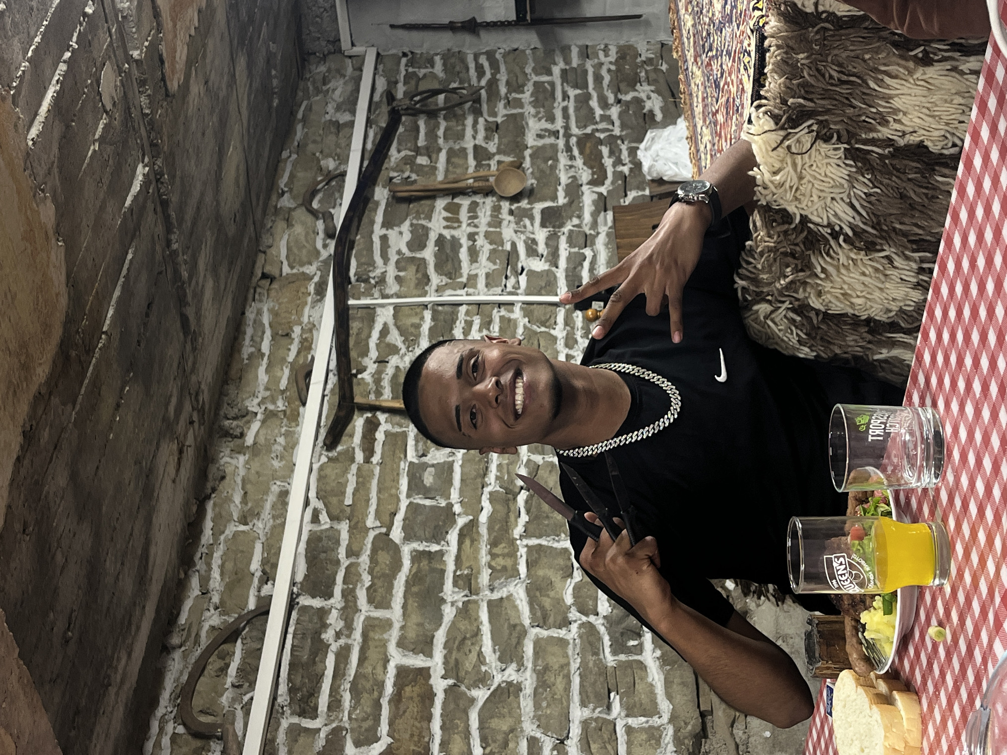 A smiling young man stands in a room with stone walls, likely within the village of Lozarevo, Bulgaria, given the image's origin. He wears a black T-shirt and a silver chain necklace. He holds two knives in his left hand, while his right hand displays a peace sign. He is seated at a table covered with a red and white checkered tablecloth. On the table, there are drinks in glasses and food items including bread and salad. The wall behind him is decorated with antique tools and implements.