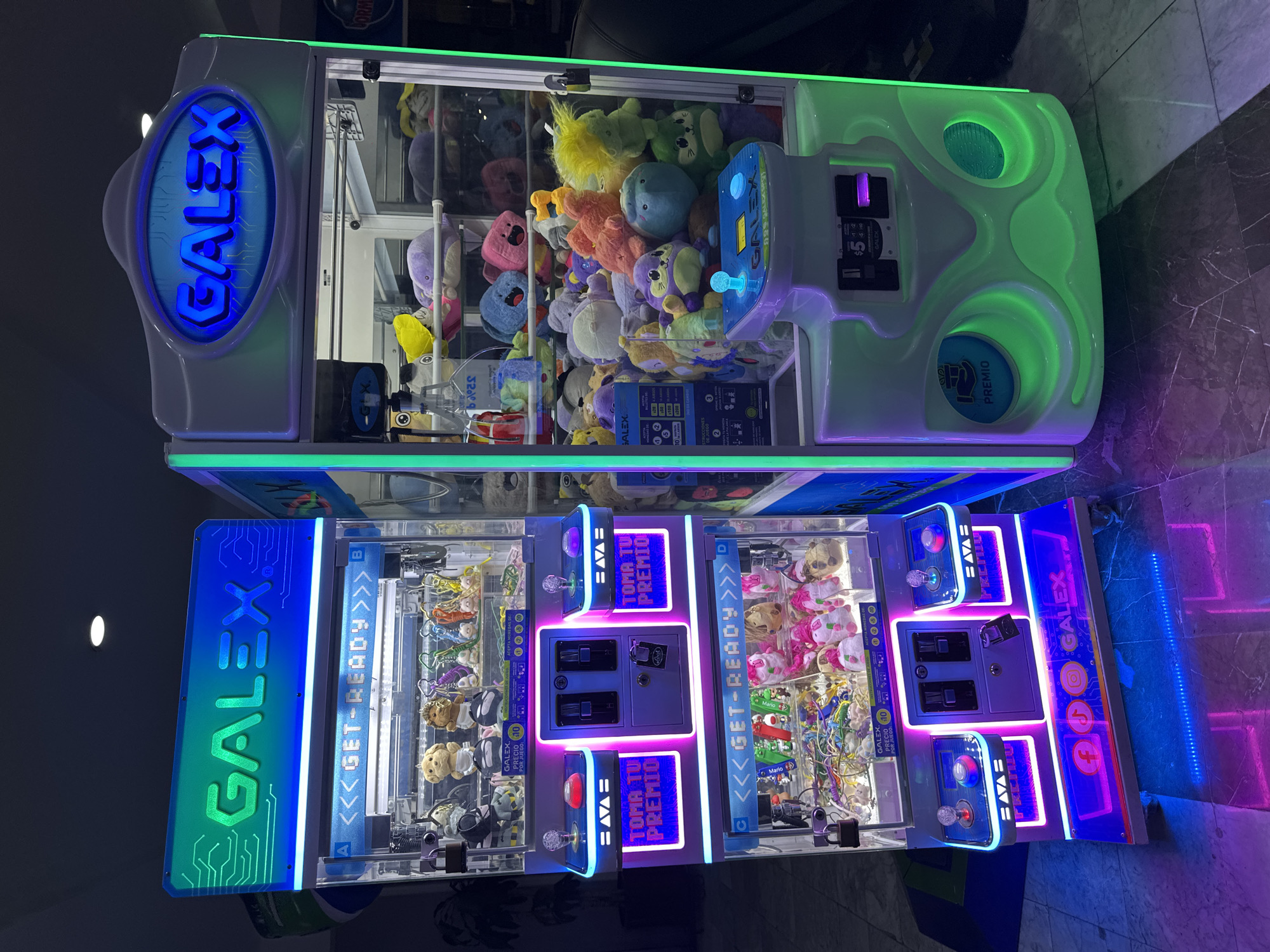 The image shows two Galex brand claw machines stacked on top of each other in what appears to be an indoor location in Huixquilucan de Degollado, Mexico. The top machine features a lime-green and white color scheme with stuffed animals visible inside. The word "GALEX" is illuminated in blue on the side. The bottom machine is two-sided, with each side being a separate claw game. The sides are predominantly blue and white, with the word "GALEX" also prominently displayed, along with the phrase "<<<GET-READY>>>". Each side has its own set of controls and a prize dispensing area. The interior of the bottom machines are filled with a variety of stuffed animals and keychains. The machines are located indoors, and the surrounding area is tiled.