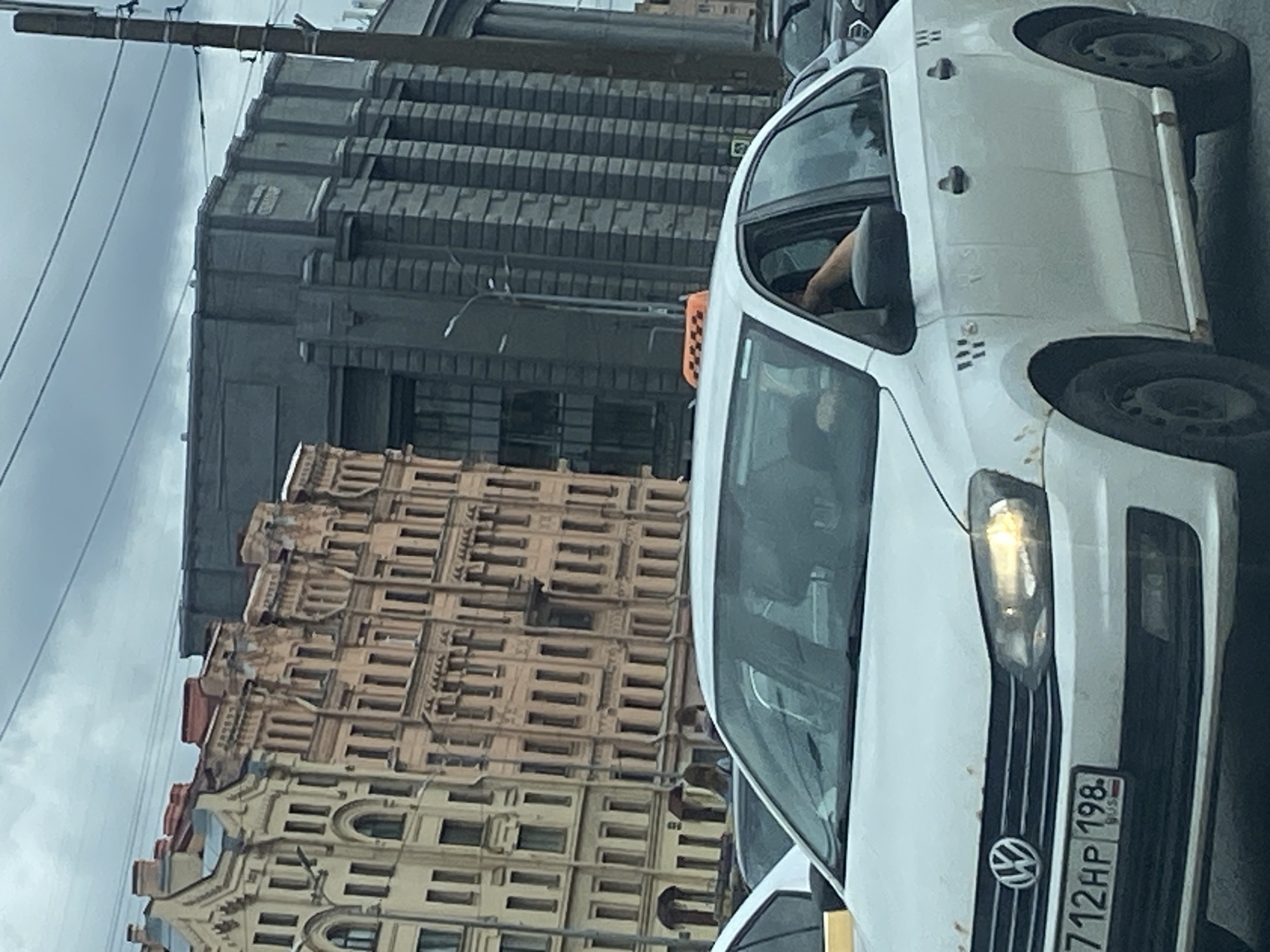 A white Volkswagen Polo taxi is parked on a street in Saint Petersburg, Russia. The taxi has a distinctive orange taxi sign on its roof. The car is facing the viewer and is slightly angled to the left. The license plate is visible and reads "712HP 198 RUS". The car appears to be weathered with some rust visible on the body. In the background, there are several buildings, including a light-colored building with intricate architectural details and a darker, more modern-looking building. The sky is overcast. A person's arm is visible through the open driver's side window.