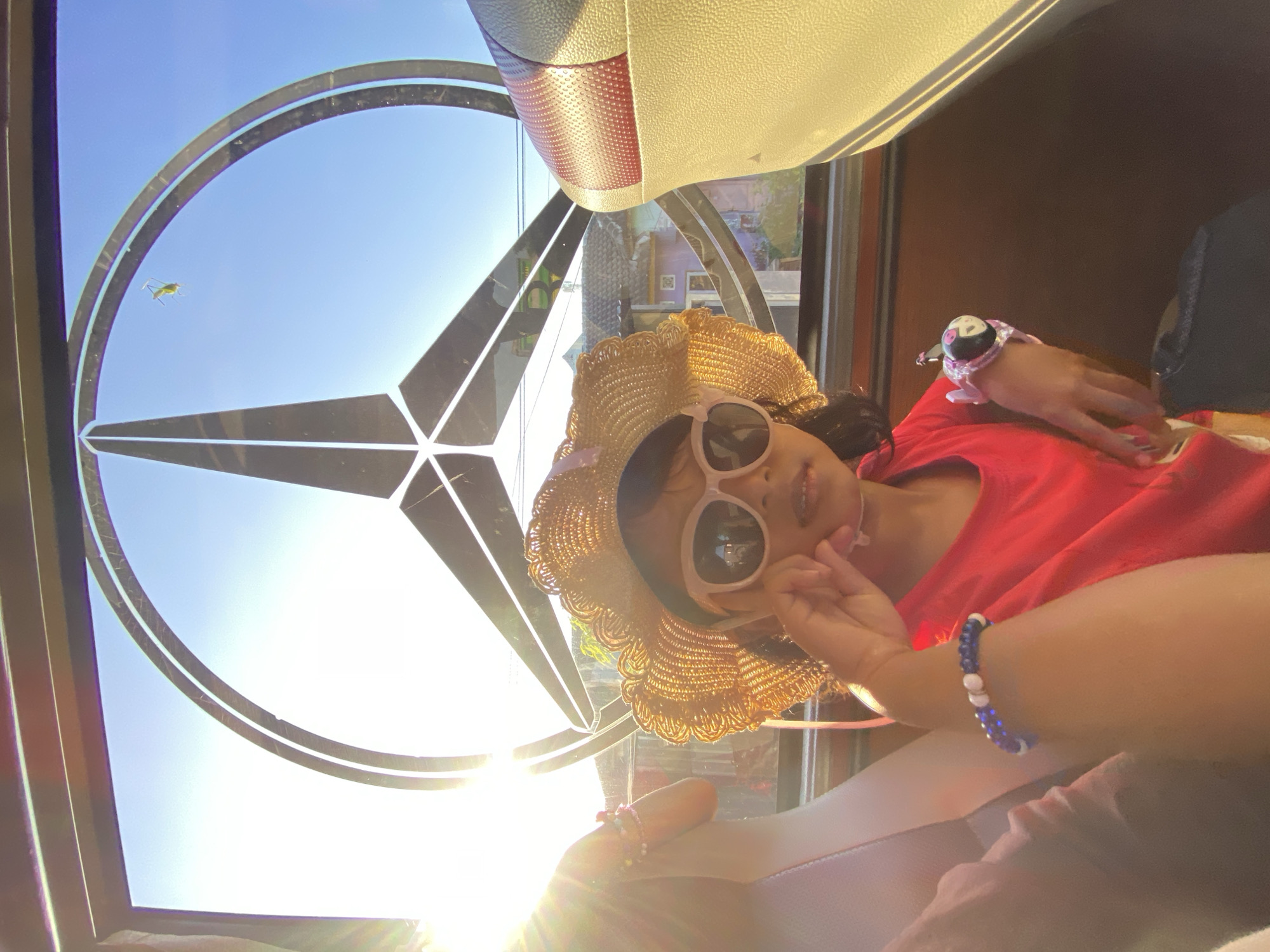 The image depicts a young child seated within a vehicle in Gelam, Indonesia, looking directly at the camera. The child wears a straw hat with a wavy brim, light pink-framed sunglasses, and a red short-sleeved shirt. A blue and white beaded bracelet is on the child's left forearm, and a pink and black wrist-mounted toy or device is on the right wrist. The child's left hand is raised near the chin, holding a thin, light-colored object. Behind the child, a large window reveals a bright blue sky and a prominent Mercedes-Benz star logo, which appears to be part of the window's structure or attached to its interior. Intense sunlight from the left creates significant lens flare across the left side of the frame. To the child's right, a beige vehicle seat with a textured red headrest is visible. On the exterior of the window, positioned within the upper right section of the Mercedes-Benz logo, a green insect is observed upside down on the glass. Through the window, distant structures with visible roofs are present in the background.