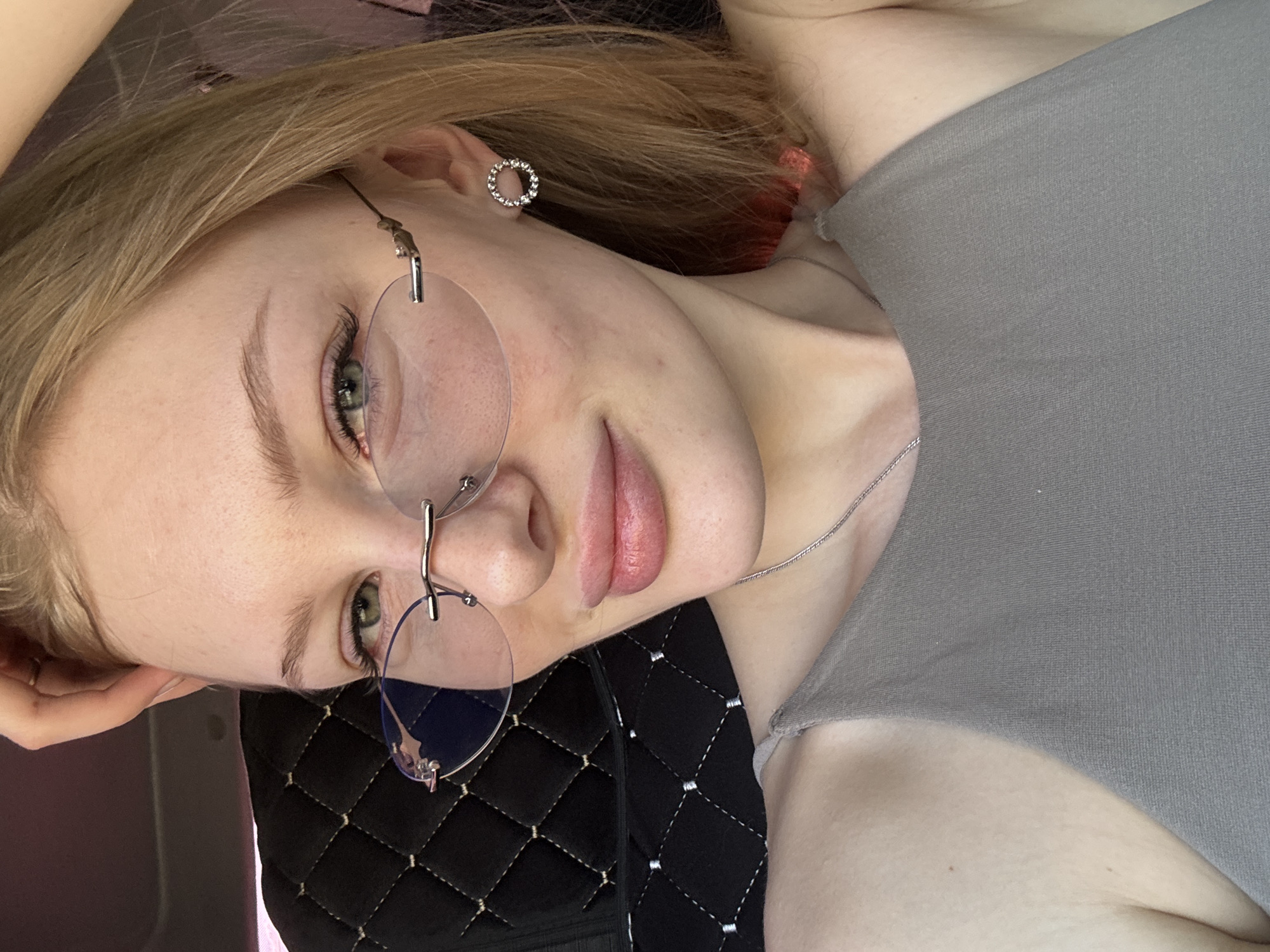 A young, fair-skinned woman with blonde hair and green eyes is angled in a close-up selfie, likely in the city of Perm, Russia. She is wearing round, rimless glasses with silver frames and a silver, circular earring in her left ear. A grey, sleeveless top is visible on her upper body. The background features a black, quilted surface, possibly a car seat. A silver necklace is around her neck. The lighting appears to be natural.