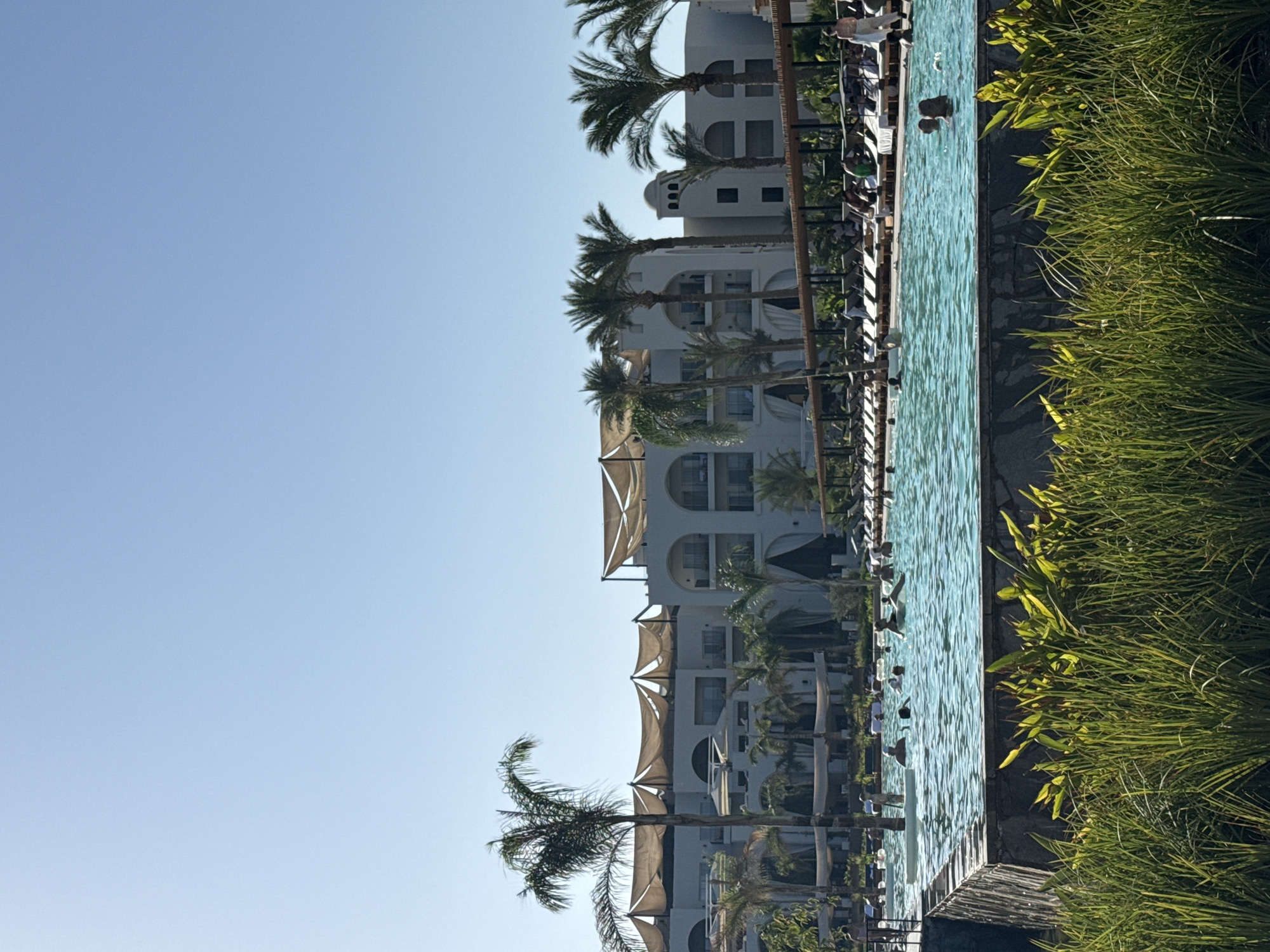 The image depicts a resort pool area in Hurghada, Egypt. In the foreground, lush green grass and foliage frame the scene. A large, clear blue swimming pool occupies the midground, with several people swimming and relaxing in the water. The pool is surrounded by a stone border, and beyond it, rows of lounge chairs with umbrellas are visible, occupied by numerous individuals basking in the sun. In the background, a multi-story resort building with arched windows and white awnings stands against a clear, bright blue sky. Palm trees are scattered throughout the resort, adding to the tropical ambiance. The overall impression is one of a sunny, recreational day at a vacation destination.