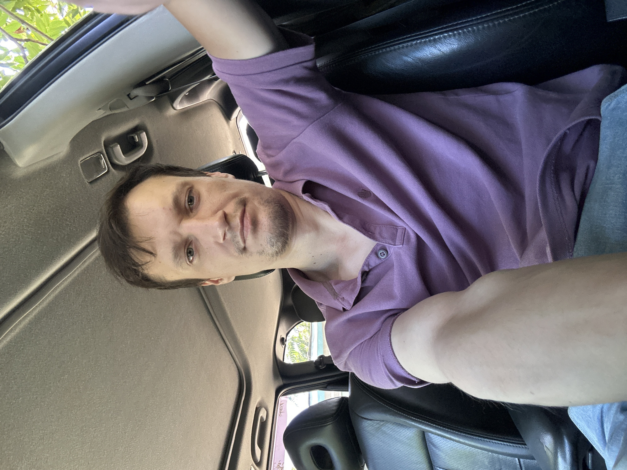 A young adult male with short brown hair and a light complexion is taking a selfie while seated in the driver's seat of a car. He is wearing a purple polo shirt. The interior of the car, including black leather seats and a sunroof, is visible. The car appears to be parked outdoors during the daytime, as indicated by sunlight filtering through the trees and a glimpse of a body of water in the background. The location is the city of Makiv, Ukraine.