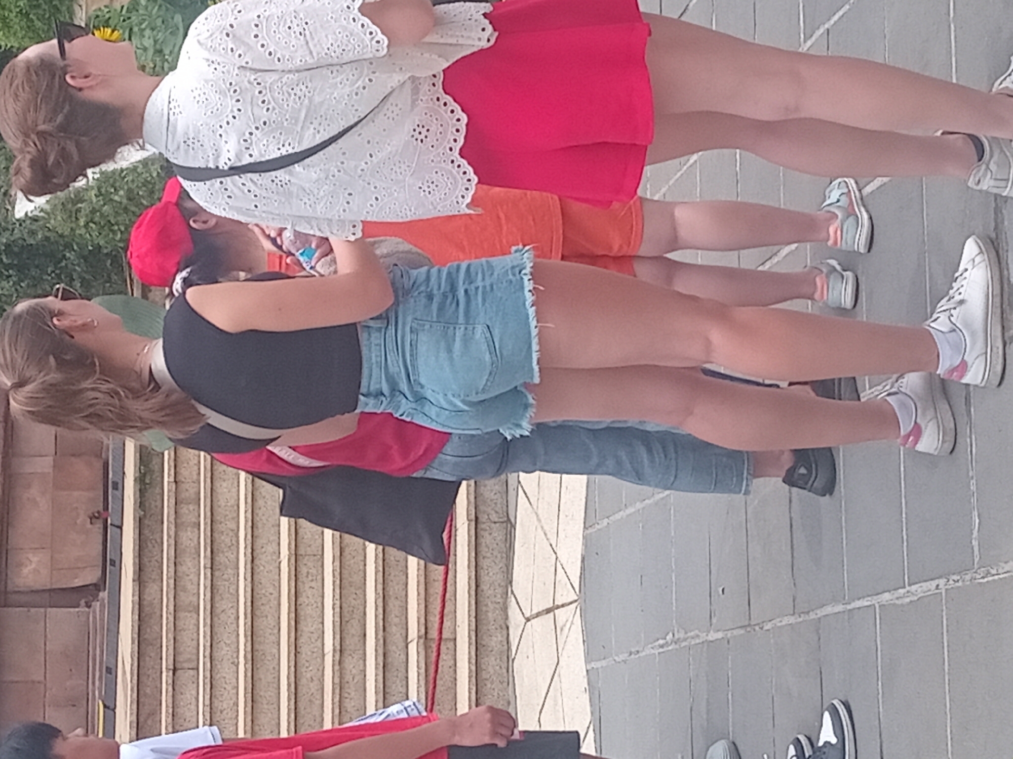 The image shows a group of people standing on a paved area in front of a set of stone stairs. The location appears to be outdoors in an urban environment, possibly a tourist attraction. The people are casually dressed, with some wearing shorts and t-shirts. One person on the right is wearing a white, lacy blouse and a bright pink skirt. Another person in the center is wearing denim shorts and a black top. A person on the left is wearing a red shirt. In the background, there are stone stairs with a red rope barrier. The overall atmosphere seems to be a relaxed, sunny day.
Without further information, it is not possible to definitively identify the specific location within Da Nang, Vietnam. However, the architecture of the stone stairs and the paved ground suggest a public area that could be found in various parts of the city, such as near a temple, a museum, or a park. The foliage visible in the background indicates a temperate or tropical climate, consistent with Da Nang.