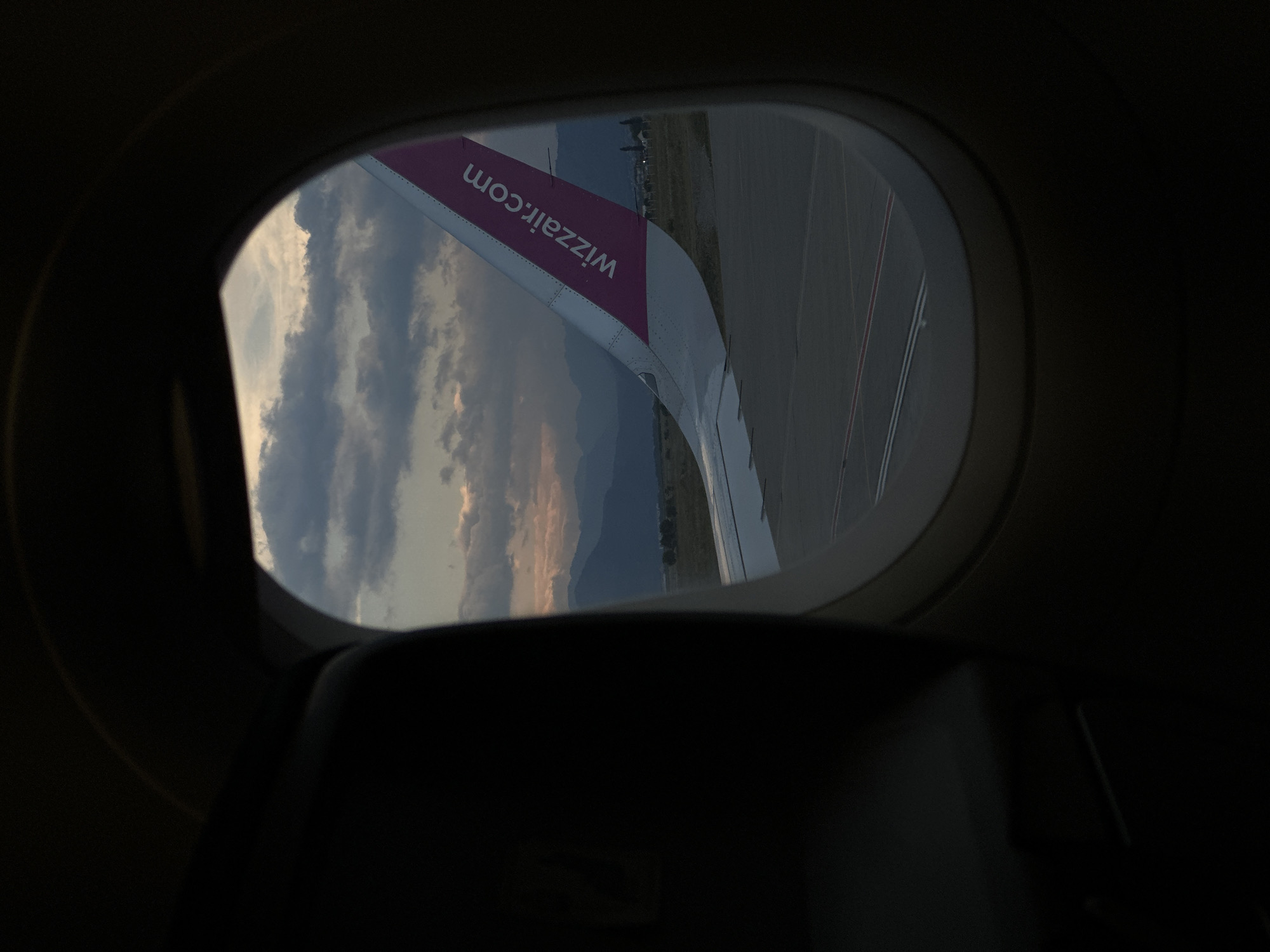 The image shows a view from inside an airplane looking out of a window. Part of the plane's wing is visible with "wizzair.com" printed on it. Beyond the wing and through the window, there are clouds, mountains, and what appears to be an airport runway or a tarmac. The lighting suggests dusk or dawn. The location, Donja Cijevna, Montenegro, may be a point of interest, perhaps the origin or destination of the flight, but no specifics are discernible from the image. The focus is mainly on the wing, sky, and airport view.