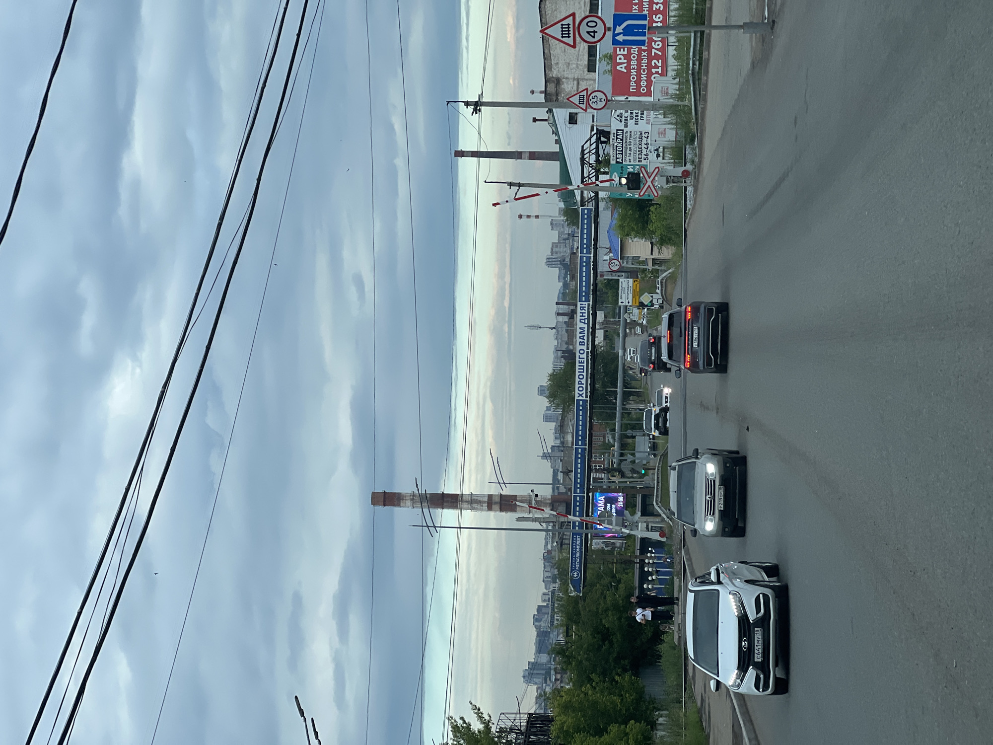 This is a vertical, eye-level, medium shot of a street scene in Izhevsk, Russia, taken from a vehicle. The weather appears overcast, contributing to a muted, diffused light, suggesting either early morning or late afternoon. The image captures a road with several cars driving on it, including a white Lada Vesta in the foreground and a silver Volkswagen Tiguan further back. The street is lined with various signs and structures, typical of a commercial or industrial area.
Prominent among these is a railway crossing with a barrier. Tall industrial smokestacks are visible in the background, hinting at an industrial zone. A blue sign with white lettering reads "ХОРОШЕГО ВАМ ДНЯ!" (Have a good day!). Other signs advertise services like "АВТОКРАН" (autocrane) with load capacities and contact information, and signage for "МЕТАЛЛКОМП" (metallcomp). Traffic signs including a "40" speed limit and a railway crossing warning are also visible. Several overhead power lines stretch across the sky, adding to the urban, slightly utilitarian feel of the scene. Two individuals, possibly pedestrians, are standing near the side of the road, observing the passing cars. The overall mood is neutral, capturing a slice of everyday life on a cloudy day in a Russian city.