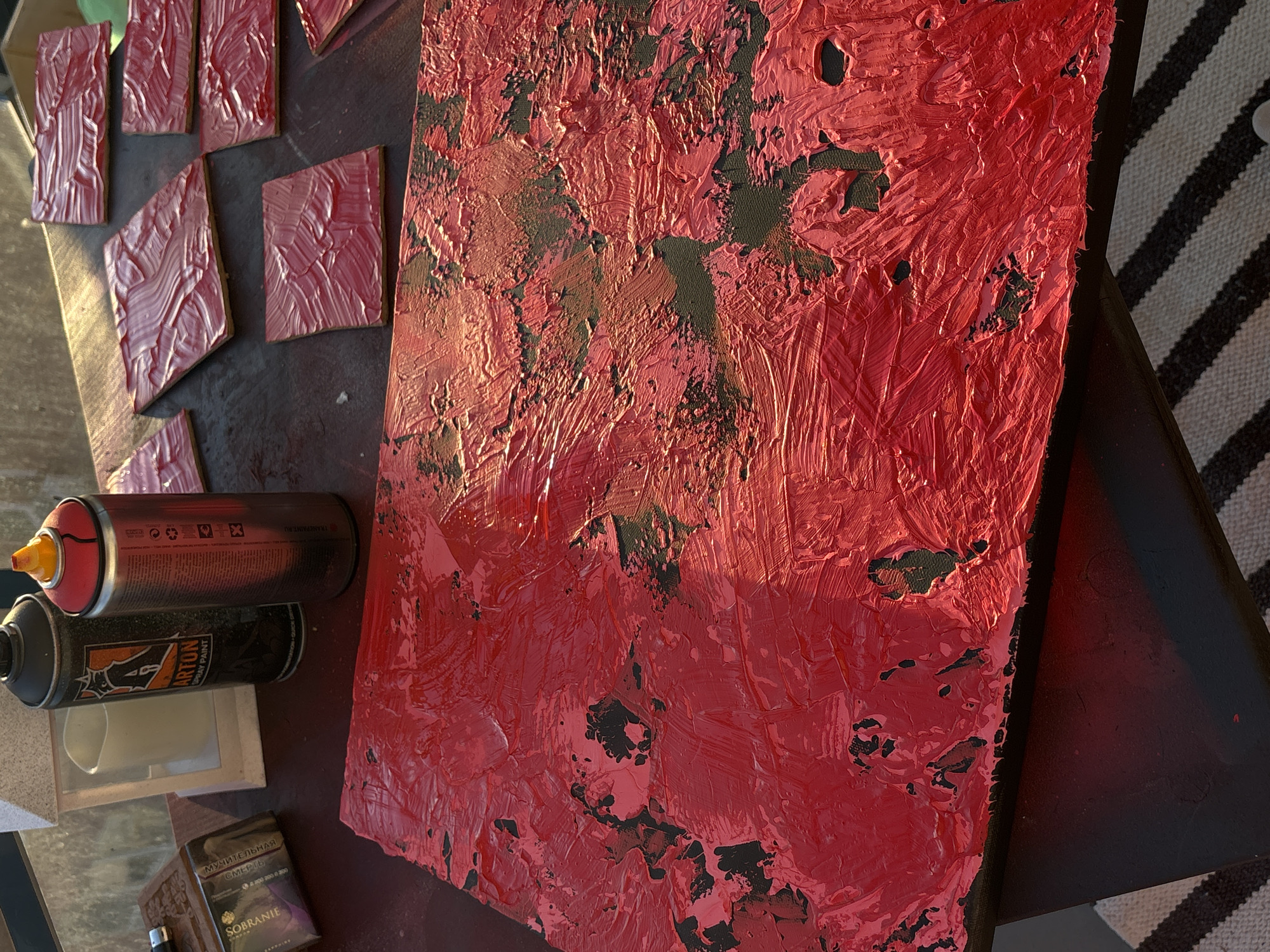 The scene shows an art project in progress, possibly outdoors given the lighting and surrounding environment. A rectangular canvas dominates the right side of the image, heavily textured with thick strokes of red paint, revealing glimpses of a dark base layer underneath. To the left of the canvas are several small, rectangular tiles also painted with a similar red texture. A can of red spray paint with an orange nozzle and a can of "Arton" spray paint lie on the surface, along with a white container and a box of Sobranie cigarettes labeled in Russian. The background includes what appears to be a striped fabric or rug and a table or surface the items rest upon. The scene doesn't provide enough information to definitively state the location as Krasnoyarsk, Russia. This would require additional visual cues such as distinctive architecture or landmarks.