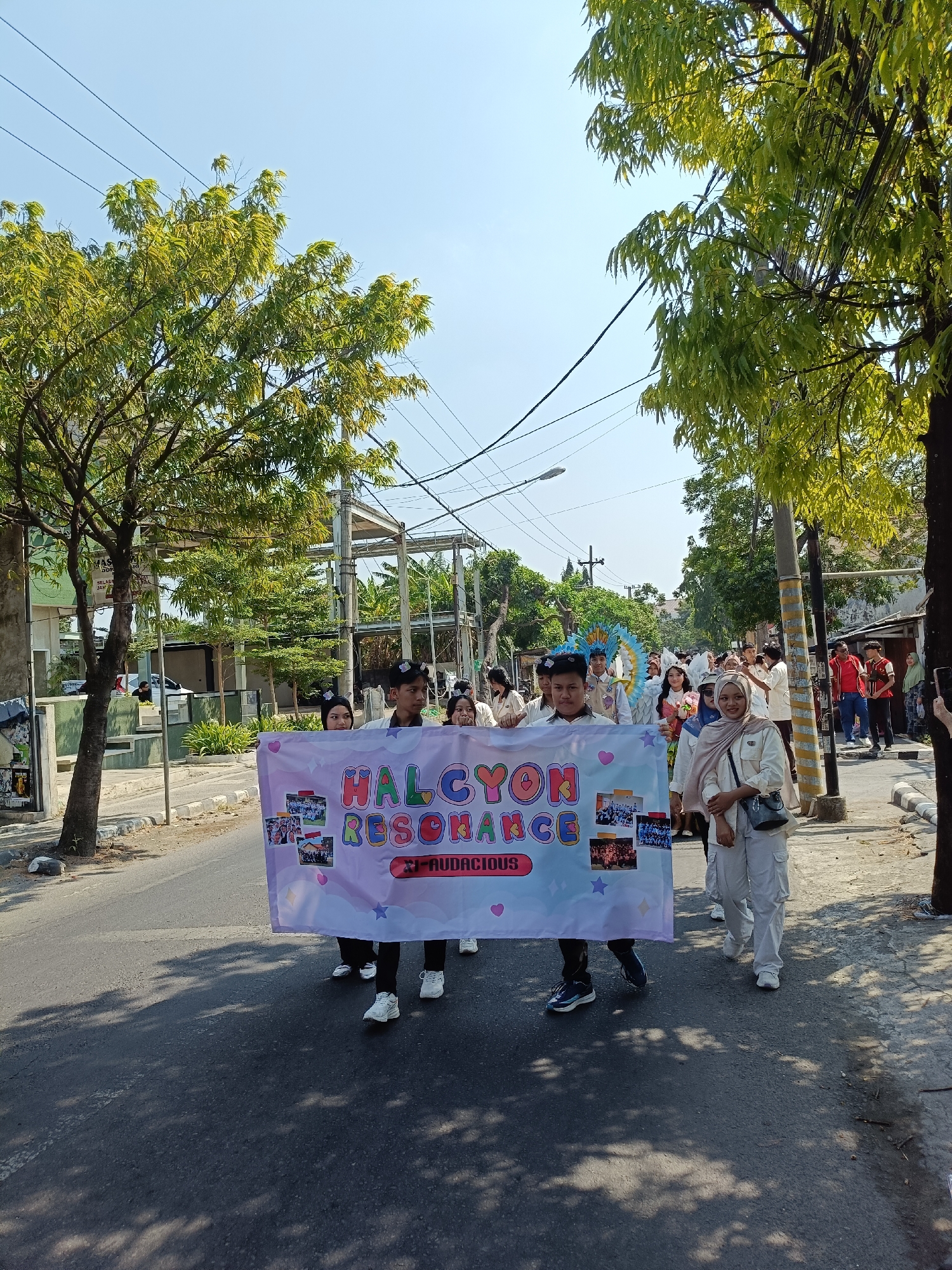 In Kediri City, Indonesia, a group of young people are participating in a parade, walking down a street. They are holding a banner with the words "HALCYON RESONANCE" printed on it, along with images. The participants are wearing various outfits, some in uniforms and others in casual attire. Some individuals have decorative headwear. The environment appears to be urban, with buildings, trees, and power lines visible along the street. The sky is clear and blue, indicating a sunny day. The people in the parade are smiling and appear to be in good spirits, suggesting a celebratory event. Some bystanders are also visible in the background, observing the procession.