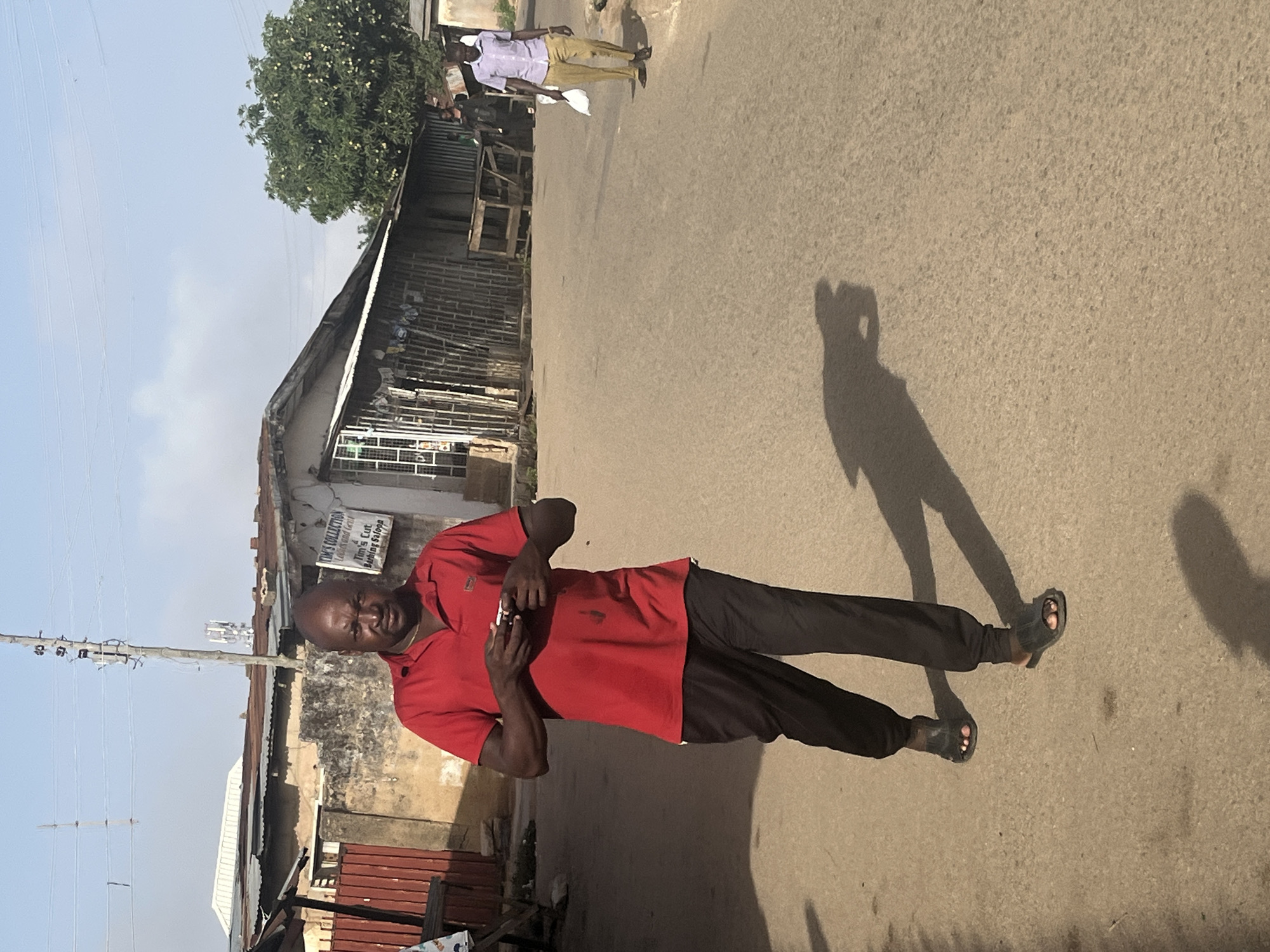 A man stands on a paved road in Jos, Nigeria. He is wearing a red polo shirt, black pants, and sandals. He appears to be holding a small object. His shadow is cast on the road to his right. Behind him is a building with a sign that reads "TIM'S COLLECTION". A utility pole is visible to the left of the building. Another man is walking in the distance further up the road. A tree is to the left, and the sky is visible.