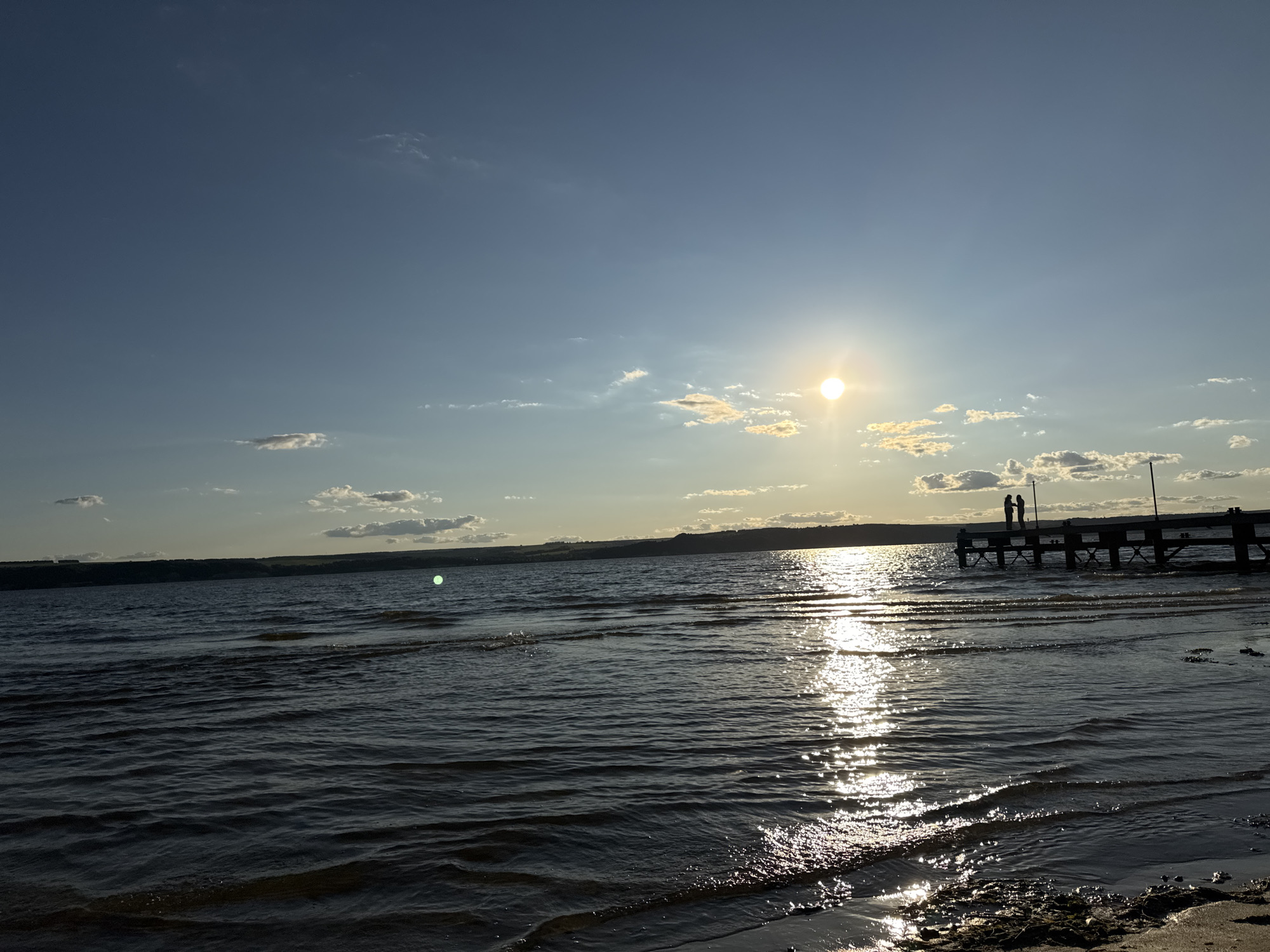 The image depicts a sunset scene over a body of water, likely a lake or river, with a pier extending into the water. The sun is positioned in the upper right quadrant of the sky, casting a strong glare on the water's surface, creating a shimmering reflection. The sky is a clear blue with scattered white clouds, tinged with yellow and orange hues from the setting sun. A silhouetted pier juts out from the right side of the image, with two small, silhouetted figures standing on it. The water is calm with gentle ripples, and the shoreline in the foreground is sandy with some debris. The background shows a low, dark horizon line of land, possibly hills or a distant treeline. The location is identified as Matyushino, Russia, suggesting this is a natural landscape within or near that area.