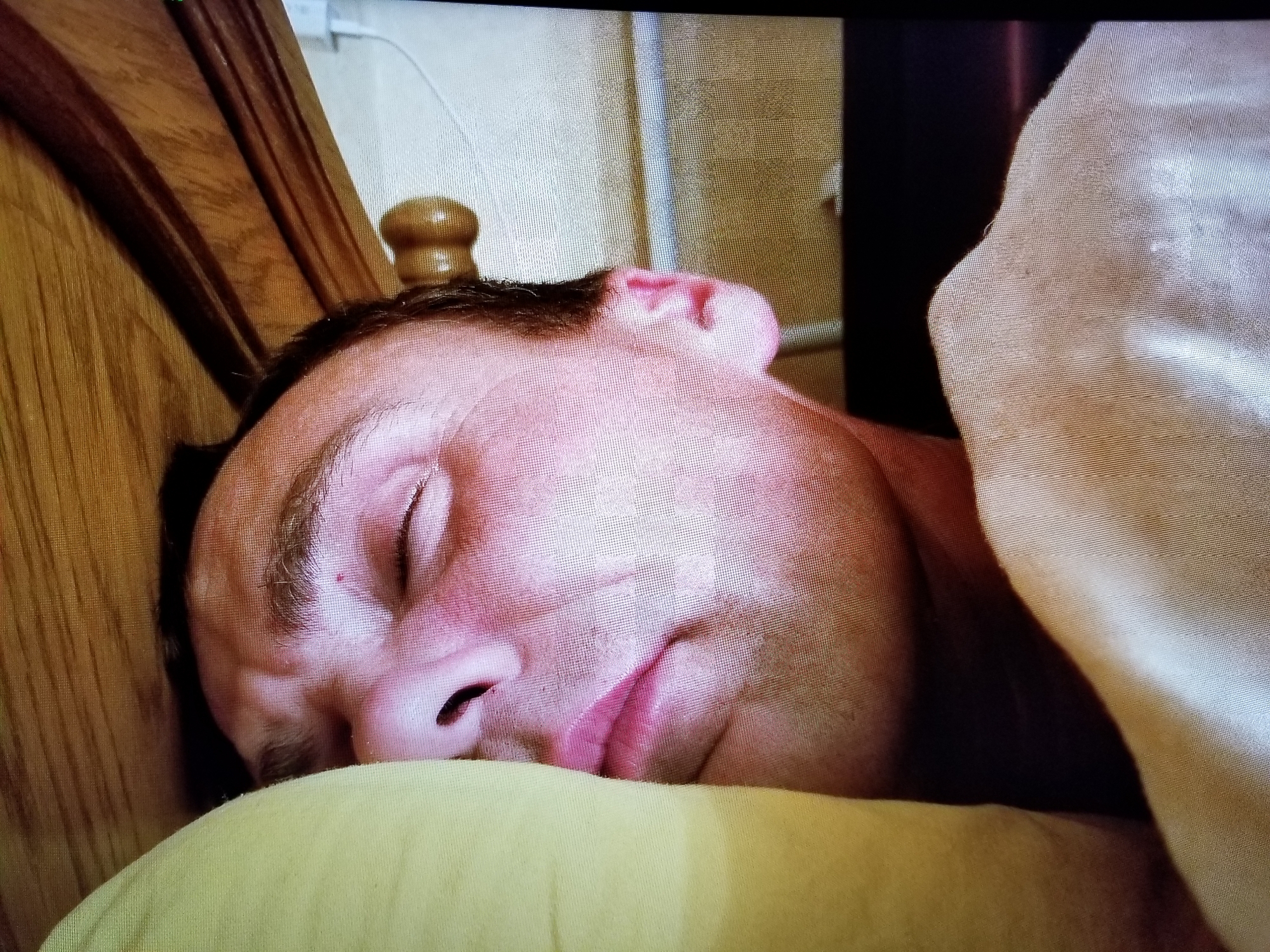 The image shows a close-up of a person sleeping on their side, with their head resting on a yellow pillow. The person's eyes are closed, and their mouth is slightly open. They are partially covered by a light-colored blanket. To the left of the person's head is a wooden headboard, and behind them is a wall with a light-colored texture. A wooden decorative knob is visible on the headboard. The scene appears to be indoors, possibly in a bedroom. There is no information in the image that would allow for the determination of its specific location within Tiraspol, Moldova.