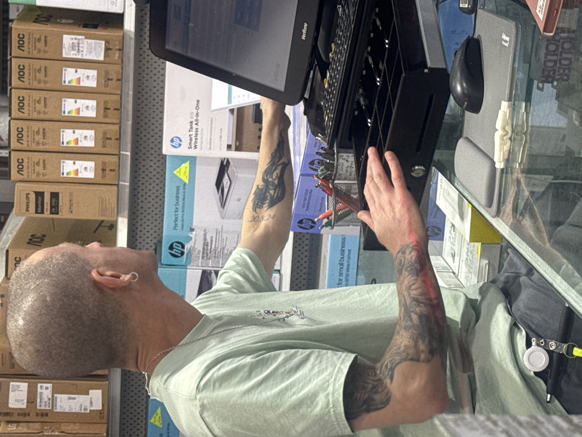 Here is a description of the image:
**Scene Description:** The image shows a man with a shaved head and tattoos on his arms standing in what appears to be a retail environment, likely a store selling electronics and office supplies. He is operating a point-of-sale (POS) system with a Verifone monitor and keyboard. The items in the store visible behind him include boxes of AOC monitors and HP printers. The countertop on which the POS system is placed appears to be clear and reflective. The man is wearing a light green t-shirt and a necklace.
**Location:** The location of Tel-Aviv is not evident in the image.