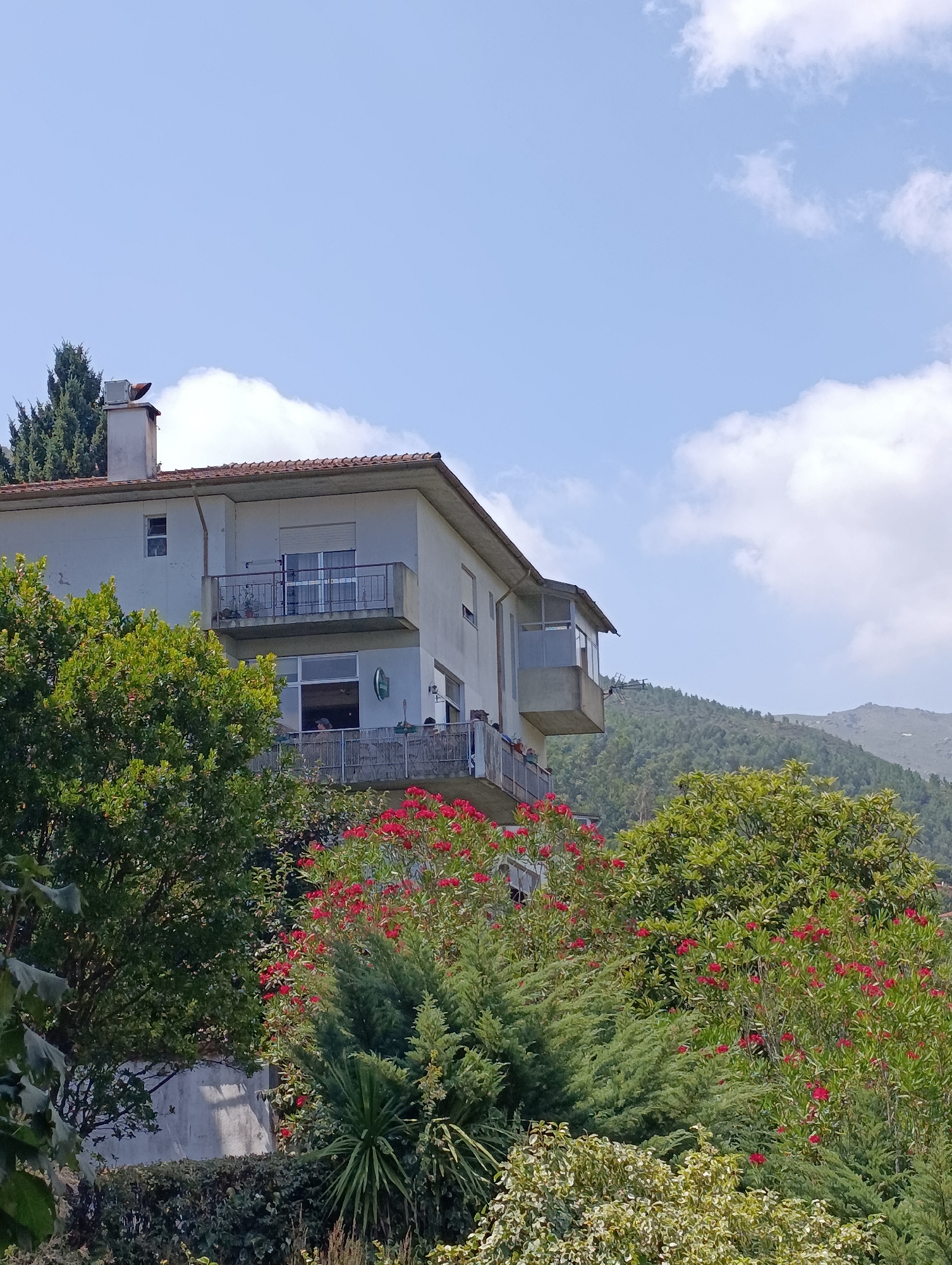 The image shows a multi-story house situated on a hillside in Terras de Bouro, Portugal. The house, painted white, has a reddish-brown tile roof and features balconies with metal railings. Red flowers and lush green foliage surround the property, with a dense forest covering the mountainous landscape in the background. The sky is blue with scattered white clouds. Two individuals are visible on the lower balcony, and several more appear to be on the upper balcony. A chimney with a bird perched on top is visible on the roof, near a tall evergreen tree. The scene suggests a residential area in a rural or semi-rural mountainous setting.