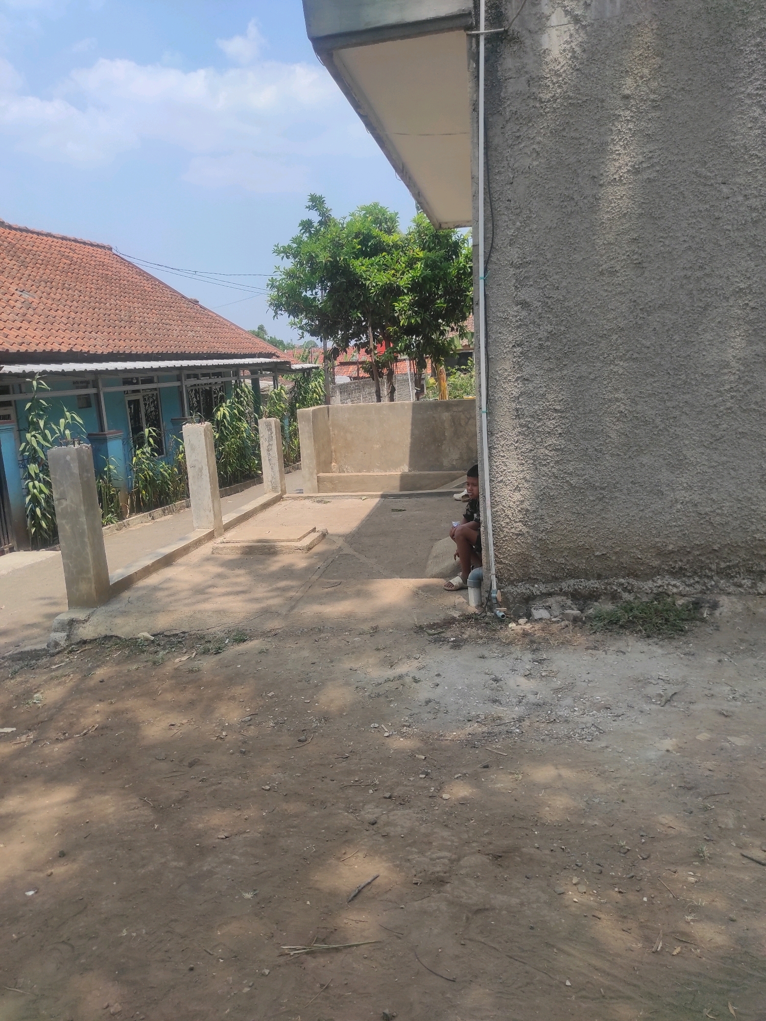 A young boy sits on a concrete ledge, partially obscured by the rough, gray wall of a building. He is wearing a dark patterned shirt and light-colored shorts, with his left leg bent and his right foot resting on the ground. A white object, possibly a cup, is held in his right hand. The building wall is textured with a rough stucco finish.
In the background, a blue house with a red tile roof is visible, surrounded by green plants. A series of low concrete barriers and posts run along a pathway leading from the house. Beyond the house, a lush green tree stands tall against a bright blue sky with scattered white clouds. The ground in the foreground is a mixture of dirt and gravel, with patches of dry grass and debris. Sunlight casts dappled shadows across the scene. The location is the city of Padalarang, Indonesia.