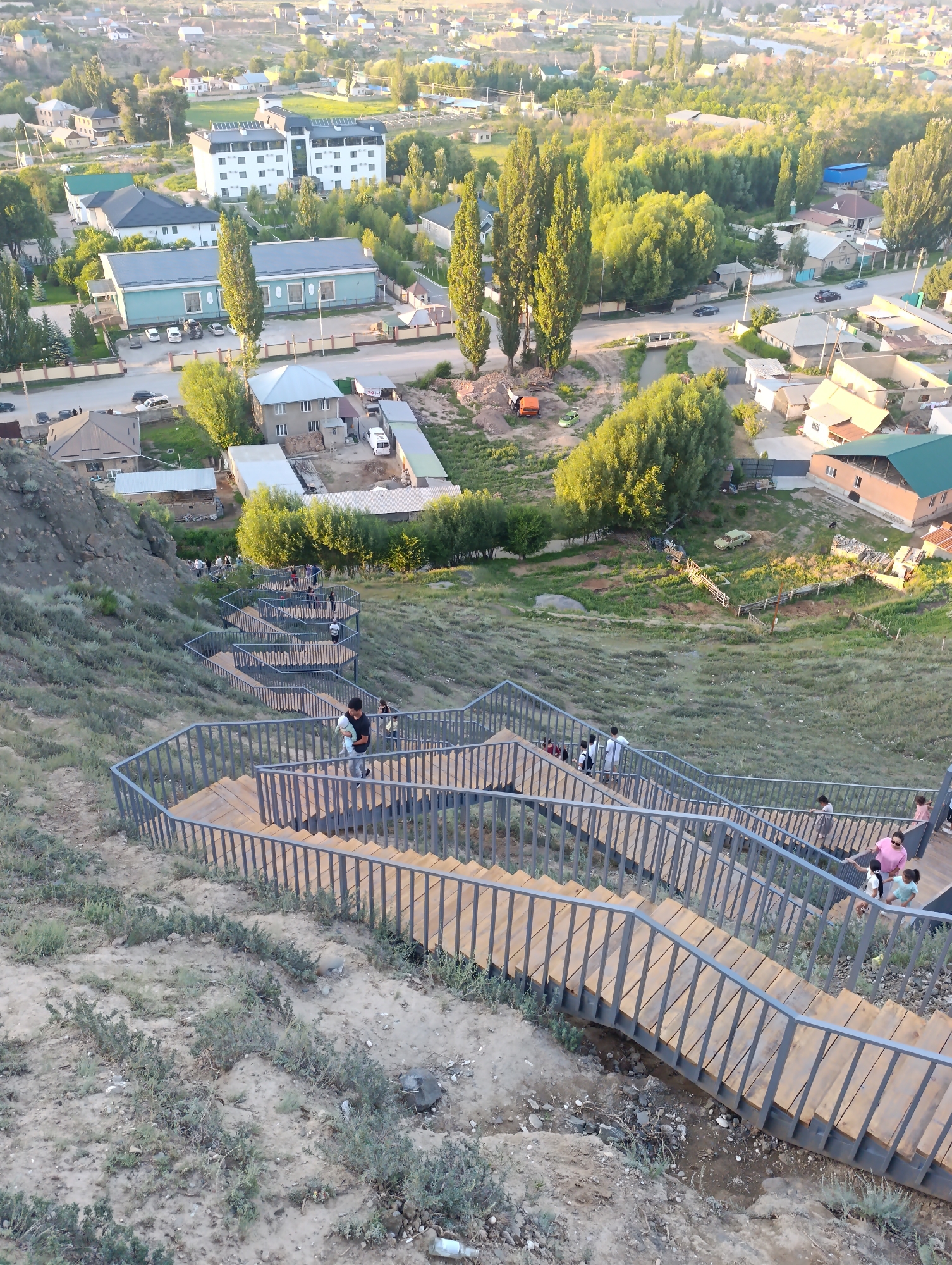 The image shows a long, winding wooden staircase with gray metal railings, descending a grassy slope. Several people are visible on the stairs, some walking down, others ascending. The stairs are a prominent feature, with a modern design and wide steps. In the background, a city or town is visible, with various buildings, trees, and streets. The architecture of the buildings suggests a residential or administrative area. The overall scene appears to be a public space, possibly a park or a recreational area, located in Naryn City, Kyrgyzstan. The lighting suggests it is either early morning or late afternoon, with the sun casting long shadows.