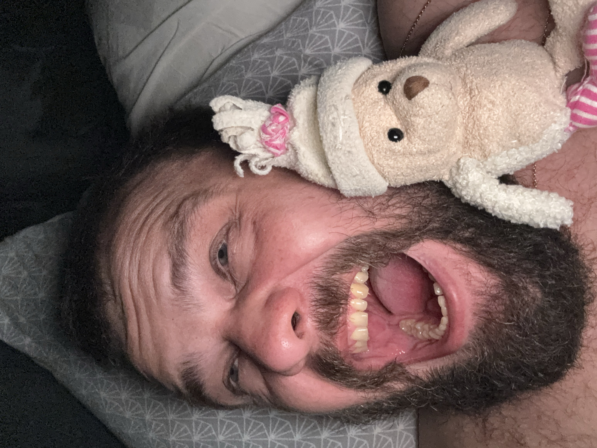 A close-up, high-angle selfie shows a person lying in bed, their face filling most of the frame. The person has fair skin, a dark beard, and is looking upwards with a wide, open-mouthed expression of surprise or excitement, revealing their teeth and the inside of their mouth. Their eyes are open and appear to be looking towards the camera.
To the right of the person's face, a small, light-colored teddy bear is held up near their ear. The teddy bear is wearing a white cap with a pink floral embellishment. It has black button eyes and a brown nose. The person is wearing a necklace.
The background consists of bedding. A pillow with a grey geometric pattern is visible behind the person's head. To the right, a light grey duvet or blanket is slightly visible. The overall lighting is dim, suggesting an indoor setting, possibly a bedroom, at night or in a dimly lit room.
There is no identifiable information or context within the image to determine the specific location in the city of Kanjoši, Montenegro.