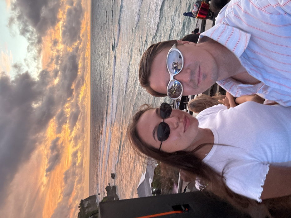 An outdoor photograph depicts a male and a female in the foreground, posing for the camera at sunset in Uluwatu, Indonesia. The female, positioned on the left, has long dark hair, wears a white t-shirt with ruffled trim, and dark oval-shaped sunglasses. To her right, the male wears a light blue button-up shirt with thin red stripes and silver, reflective, futuristic-style sunglasses. Both individuals are looking directly at the camera with slight smiles.
The background features a vast ocean under a sky displaying a sunset with prominent orange, yellow, and gray cloud formations. Waves are visible on the ocean's surface, reflecting the sky's hues. On the far left, a lush green cliff face with rock formations descends towards the water. A large, dark rectangular speaker cabinet is partially visible in the left mid-ground. Indistinct figures of other individuals are discernible in the blurred background, suggesting a public outdoor venue or event space situated on a cliff overlooking the ocean.