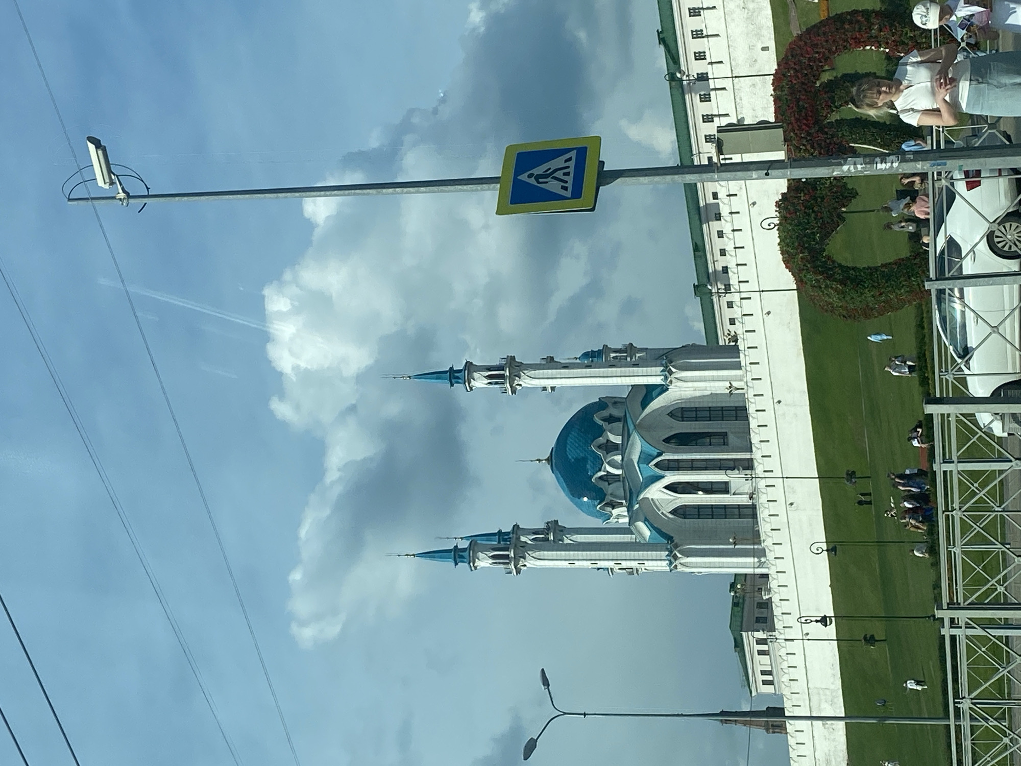 In Kazan, Russia, the Kul Sharif Mosque stands prominently against a cloudy sky. The mosque features a large blue dome and two tall white minarets with blue accents. A white wall with a green roof runs along the base of the mosque. In the foreground, a pedestrian crossing sign is mounted on a pole, and a woman in a white shirt and blue skirt is standing next to it. Several people are visible on the grassy slope in front of the mosque wall, some sitting and others standing. A white car is parked in front of a metal fence in the lower right portion of the image. The overall scene depicts a public area with a significant architectural landmark.