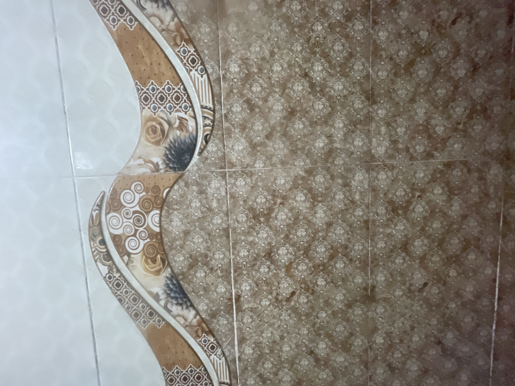 The image shows a close-up view of a tiled wall, likely in a bathroom or kitchen, in Masazir, Azerbaijan. The wall is divided horizontally by a decorative border. The upper section of the wall features a patterned tile with a wavy brown and white design that incorporates floral and geometric motifs. The lower section of the wall consists of smaller, square tiles with a repeating floral pattern in shades of brown and cream. The tiles appear to be clean and well-maintained. There are no people or other objects visible in the frame. The lighting suggests an indoor environment, possibly with artificial light.
