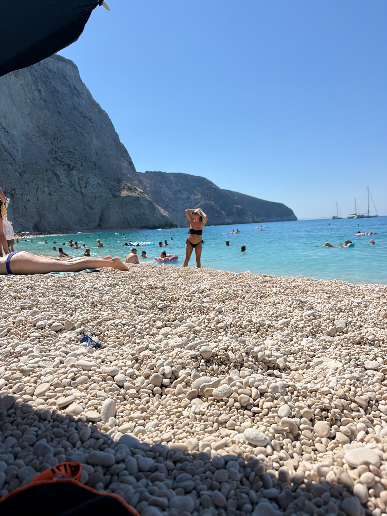An eye-level, wide shot captures a sunny day at a pebble beach in the Municipal Unit of Apollonioi, Greece. In the foreground, the legs of a person are stretched out on the beach, with their feet visible and toes pointed upwards. The beach is covered in smooth, light-colored pebbles. A dark blue object, possibly a piece of trash, lies on the pebbles near the center of the frame. In the middle ground, the turquoise waters of the Ionian Sea are populated with numerous people swimming and floating on inflatable tubes. A woman in a black bikini stands facing away from the camera, with her hands on her head, posing. Further out in the sea, sailboats are visible on the horizon. The background features a dramatic, steep, gray cliff face that curves along the coastline, with the clear blue sky above.