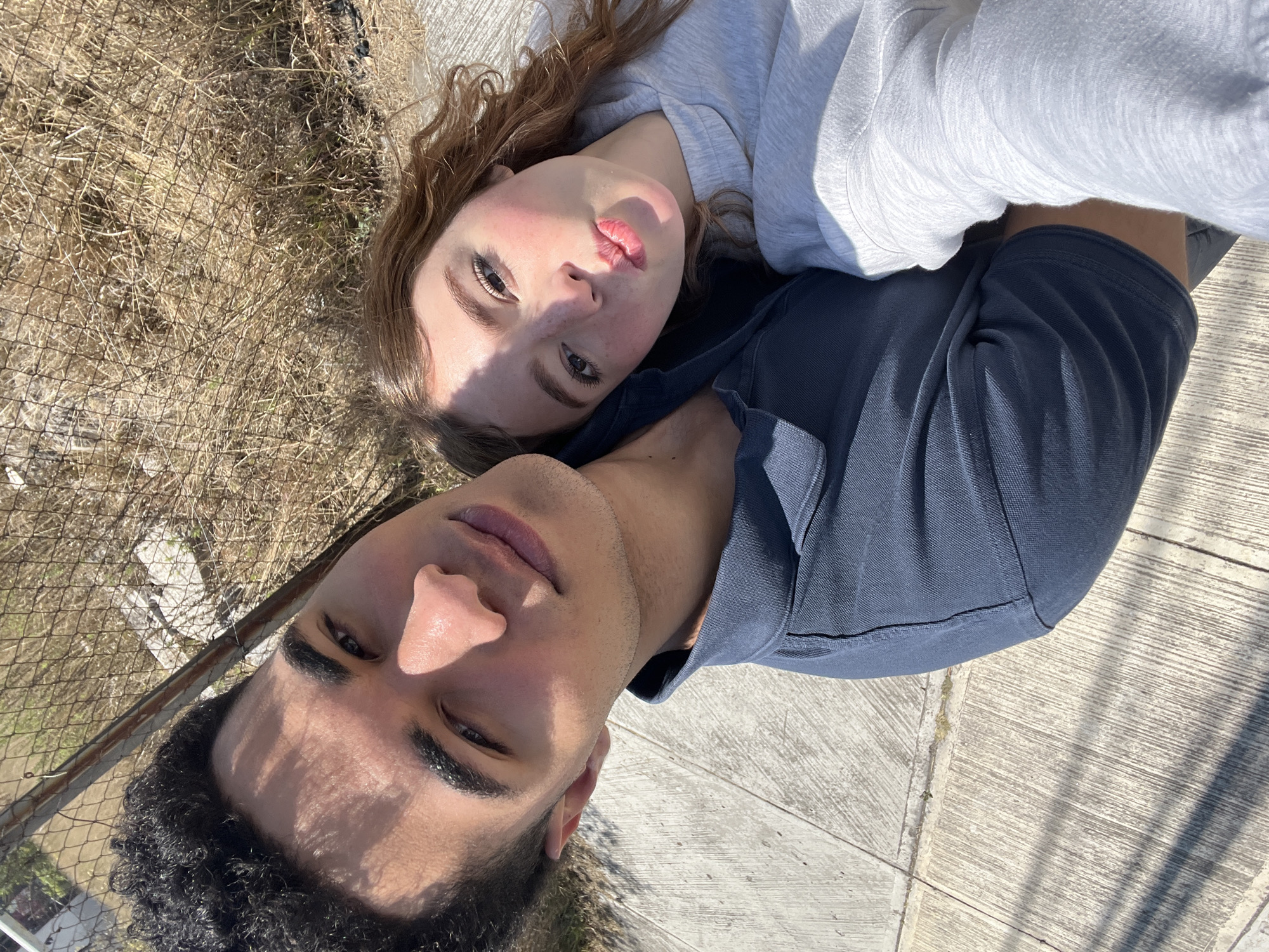 Two individuals are lying on a concrete surface next to a chain-link fence and dried vegetation. The person closest to the viewer has dark curly hair and is wearing a navy blue shirt. The other person has light skin and reddish-brown curly hair, wearing a light grey top. Both are looking up toward the camera. The location may be San Bernardino Tlaxcalancingo, Mexico.