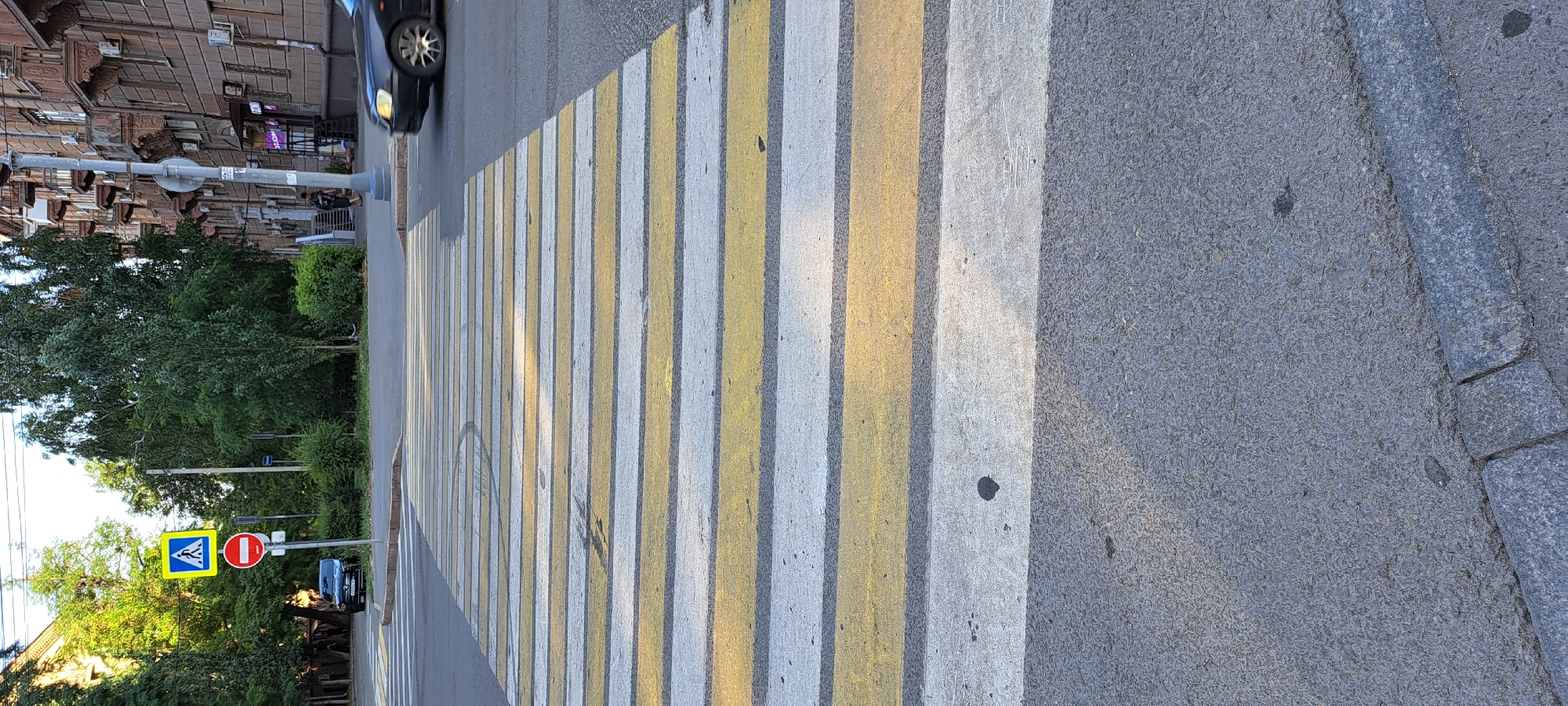 A vertical, eye-level shot shows a pedestrian crossing at a city intersection. The crosswalk, marked with alternating white and yellow stripes, dominates the foreground and extends towards the background. Beyond the crosswalk, a black car is visible in the distance, partially obscured by trees. To the left of the crosswalk, a black car is parked near the curb, alongside a pedestrian crossing sign and a circular red and white prohibition sign. Trees line the street on both sides, and an apartment building with balconies is visible on the right. The pavement is asphalt, with a visible curb at the bottom of the frame. The scene is illuminated by natural daylight.