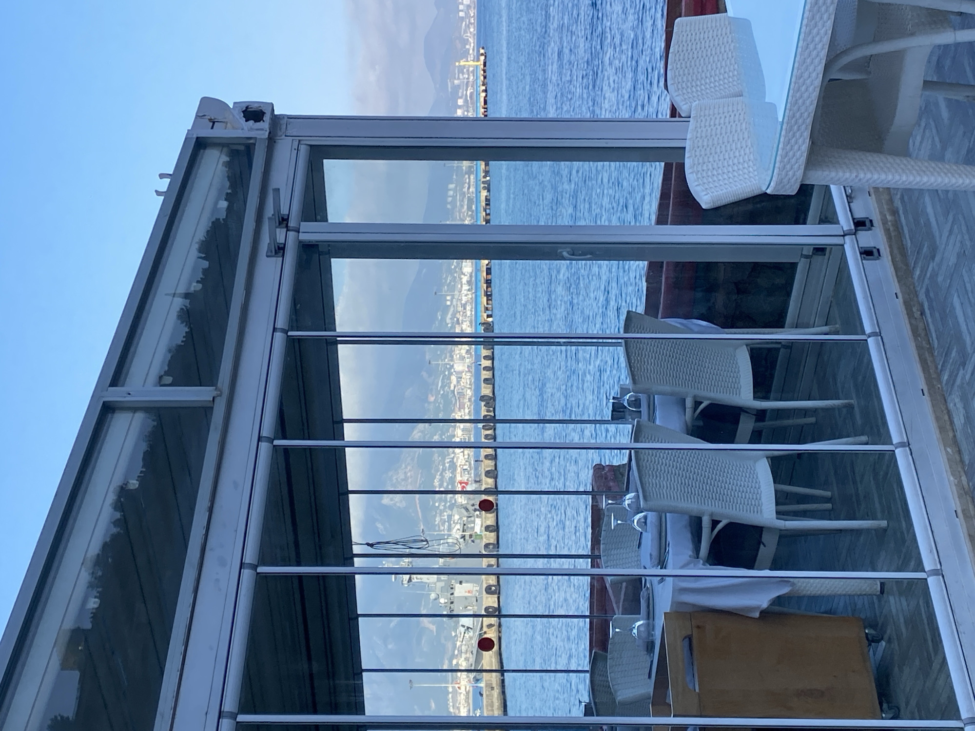 This image, when viewed in its correct orientation, captures a serene daytime scene from within a glass-enclosed dining or seating area overlooking a tranquil blue sea. The setting appears to be a restaurant or cafe, possibly along a waterfront.
In the foreground, the modern structure features a metal frame supporting large glass panels that offer unobstructed views. These panels create strong reflections of the sky and distant landscape. Inside the enclosure, several white wicker or rattan chairs are arranged around white tables, some topped with glass. White tablecloths are draped on some tables, and clear wine or water glasses are placed upside down, indicating the area is set and ready for guests. A light wooden serving cart or box on wheels is visible near one of the tables.
Beyond the glass, the calm, bright blue sea dominates the mid-ground. A long pier or breakwater, dotted with dark bollards and two noticeable red circular markers, stretches across the water. On the opposite shore, a dense cityscape with numerous buildings is visible, backed by imposing mountains under a clear, pale blue sky. A yellow industrial crane can be spotted in the distant port area.
The overall impression is one of peacefulness and an inviting atmosphere. There are no people present, suggesting either an early morning, late evening, or a quiet period. The weather is clear and sunny. No discernible text is visible within the image. This coastal setting aligns with the provided context of Alanya, Turkey.