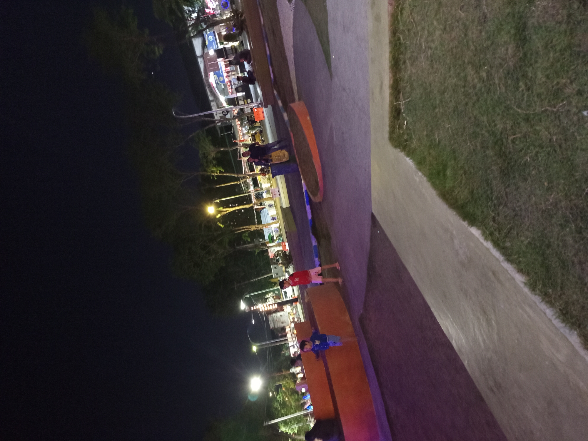 The image captures an outdoor scene at night, likely a public area or park in Sidoarjo, Indonesia. In the foreground, a concrete path with a grassy edge runs diagonally across the frame, transitioning to a dark purple textured surface. Further back, a raised, orange-colored structure is visible.
Two young boys are standing near this structure. One, wearing a blue shirt and shorts, is leaning on the orange surface. The other, in a red shirt and light-colored shorts, stands to the right.
Beyond the boys, a paved area features several people sitting or standing. A blue trash can is also present. In the background, trees line the street, illuminated by streetlights. Buildings and a vehicle are visible further back, suggesting a commercial or public establishment. The overall impression is of a well-lit urban space after dark.