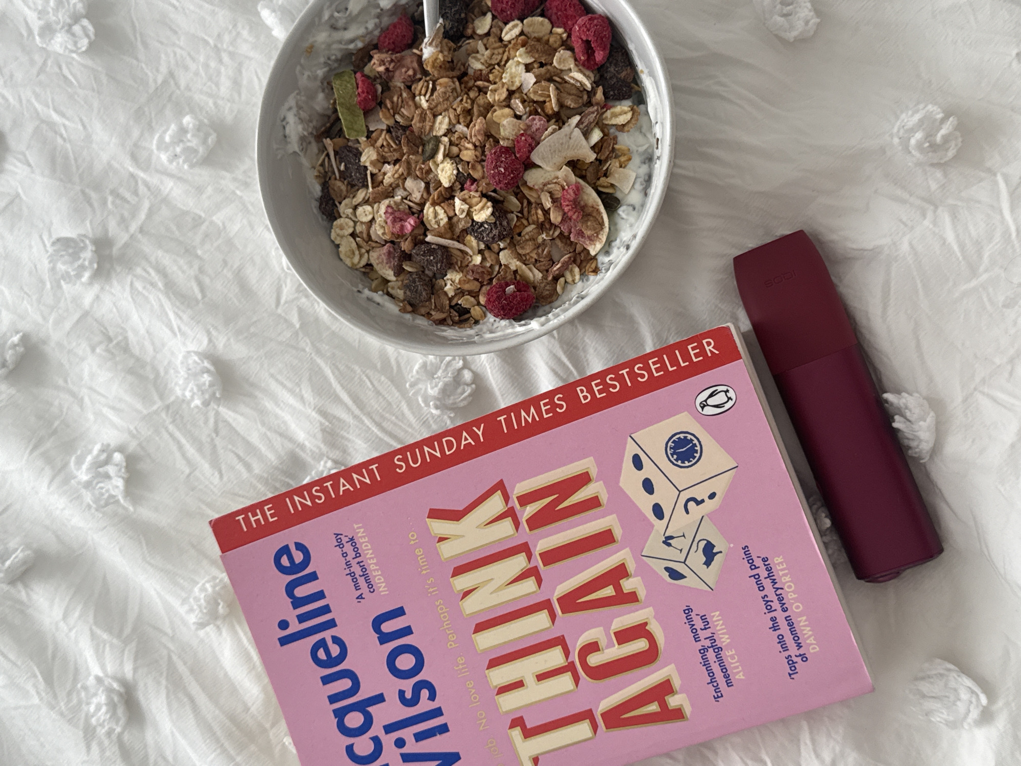 The image shows a bowl of granola and yogurt next to a book and a small, maroon-colored object on a white textured surface. The book is titled "Think Again" by Jacqueline Wilson. The maroon object is unidentifiable, but has the word "SODI" written on it. The environment appears to be a bed or a similar soft surface. There is no information to suggest this image was taken in London, United Kingdom.